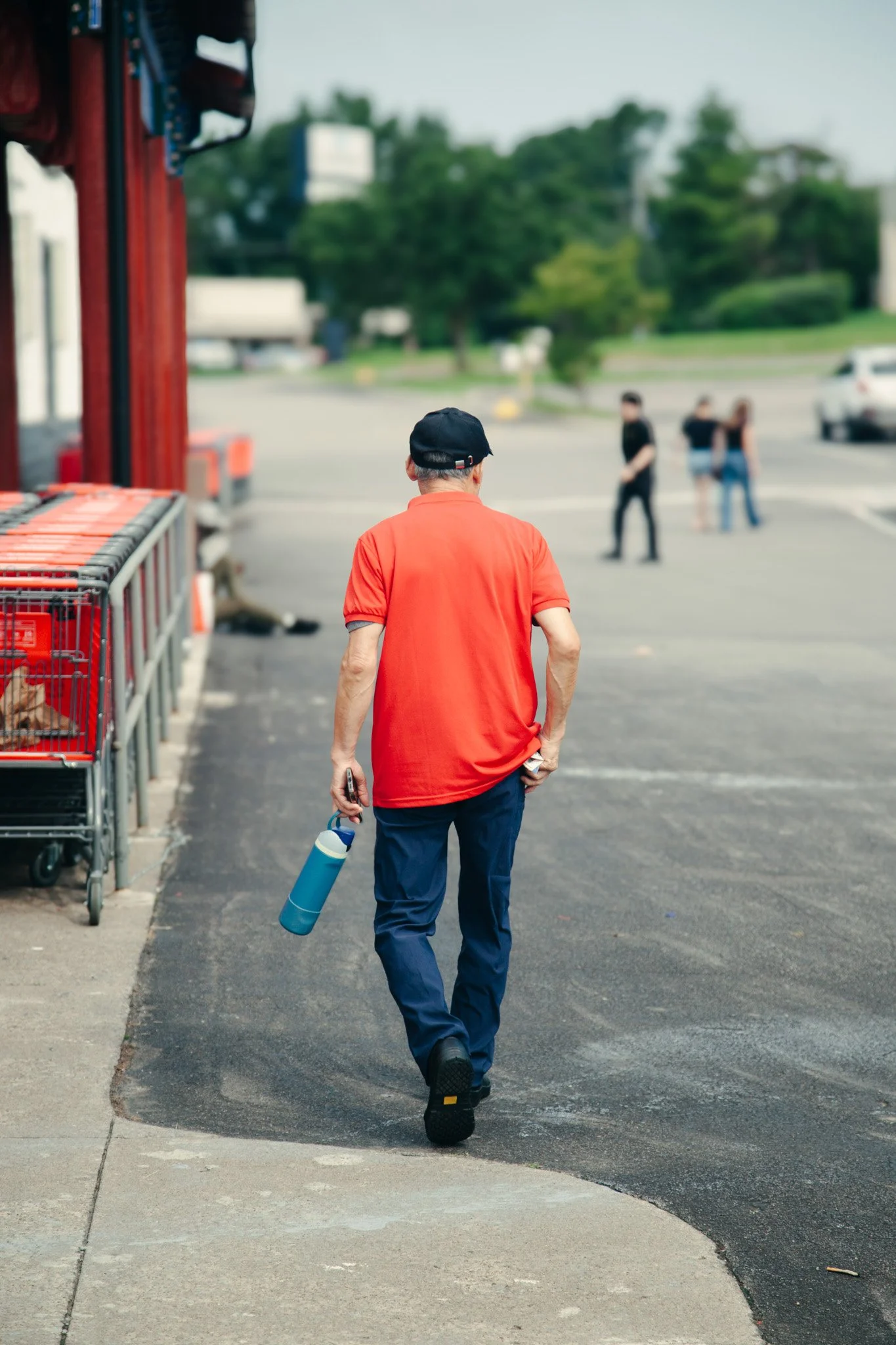 A man wearing a red shirt, black cap, and blue pants walks away from the camera, holding a blue water bottle, in an outdoor parking lot. In the background, a few people are walking and talking near parked cars, with trees and buildings further back.