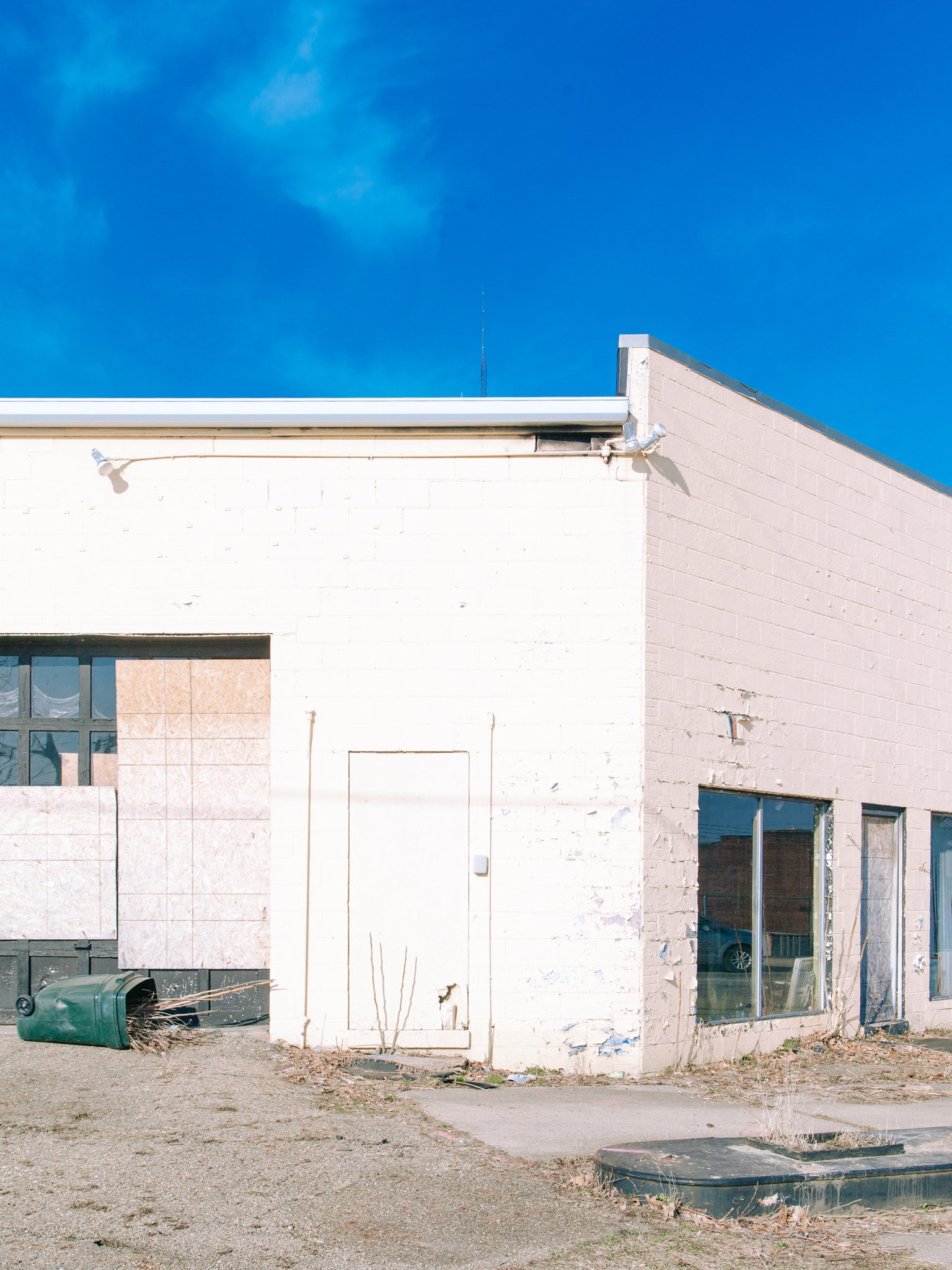 A white commercial building with boarded-up windows and a garage door, with a houseplant and debris outside, and a bright blue sky above.