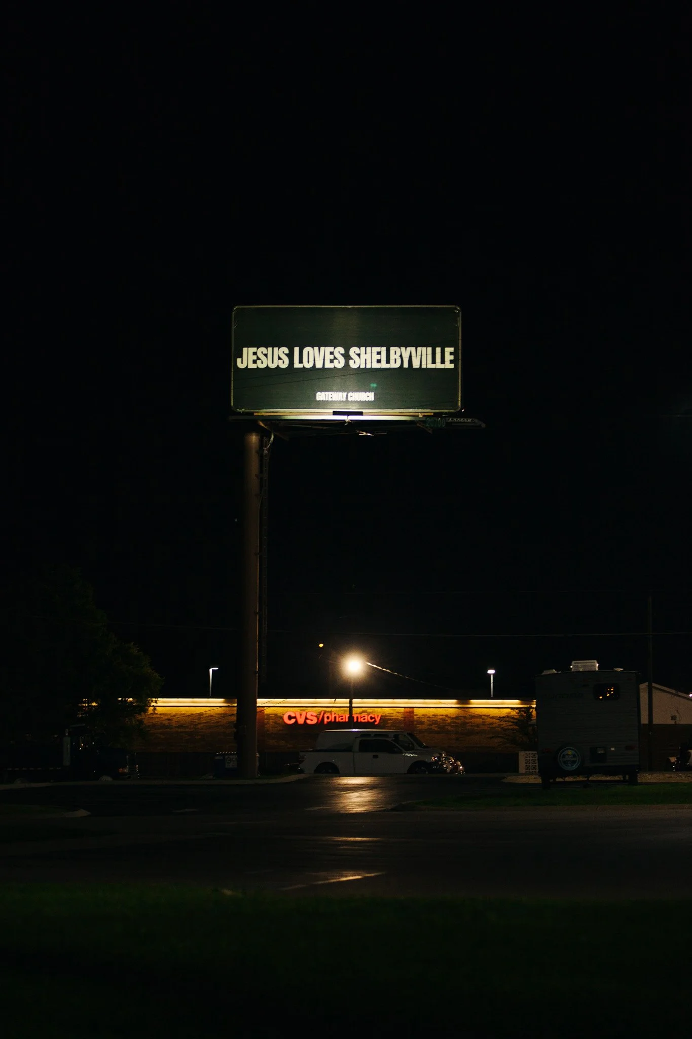 Nighttime view of a billboard that reads "JESUS LOVES SHELBYVILLE" with a smaller sign below indicating "GATEWAY CHURCH". Below the billboard, a CVS pharmacy is visible with its brightly lit sign. There are some vehicles and streetlights along the ro