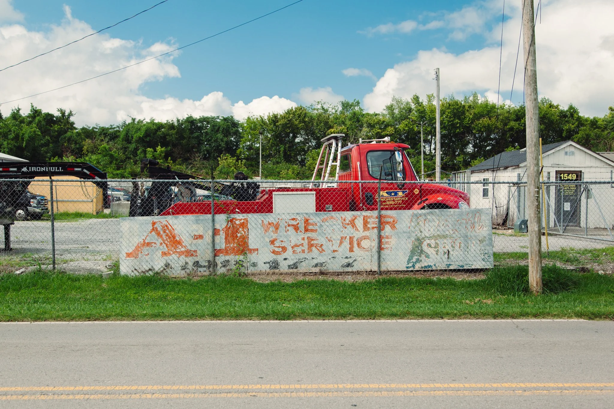 A red service truck with a crane parked behind a chain-link fence. There is a sign on the fence that is partially visible, indicating it is a wire transfer service, and a house with a gray roof and a house number 1549 in the background. The scene is 
