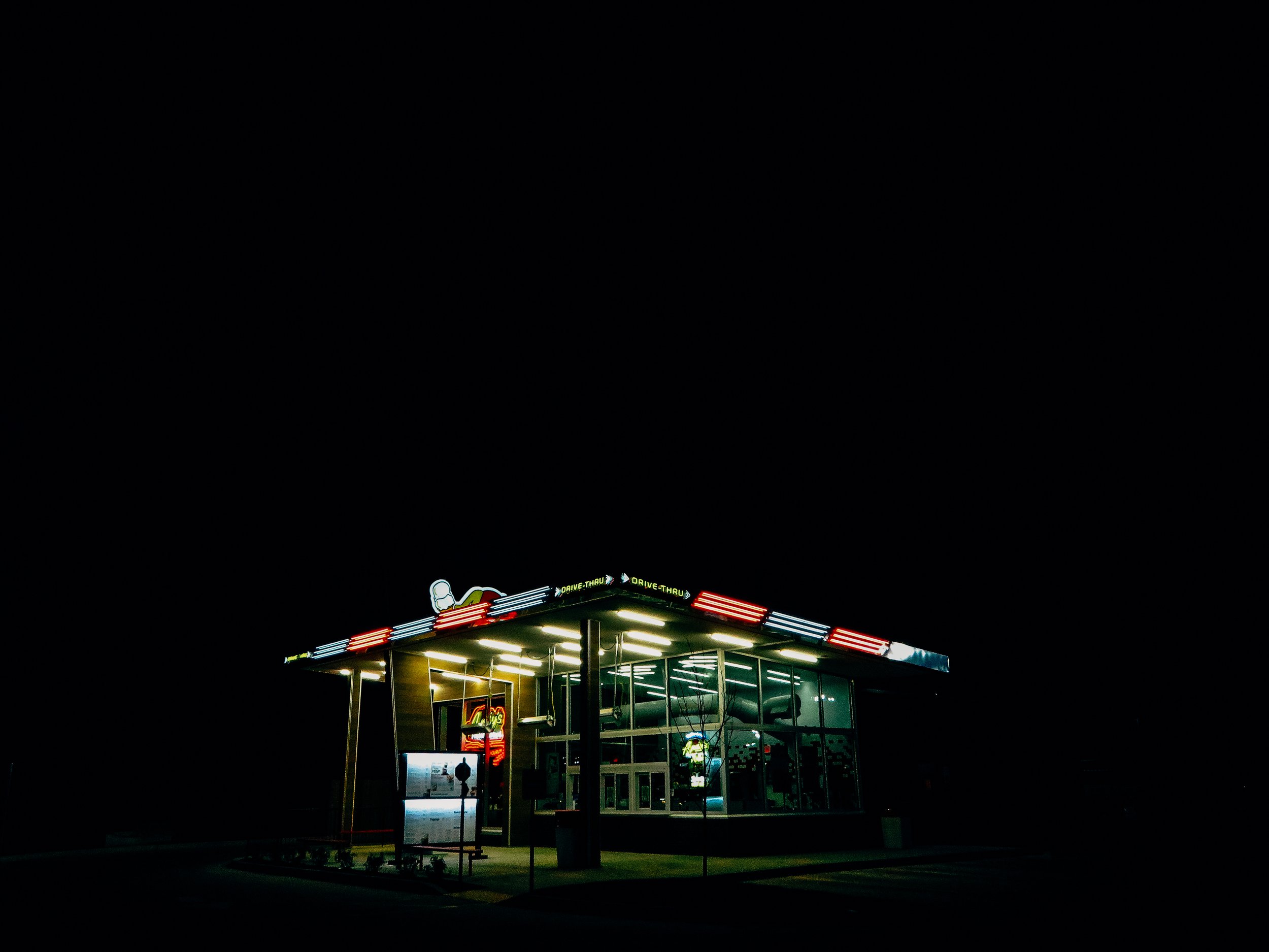 Night view of a modern building with illuminated neon signs, including drive-thru signs, with a dark sky background.