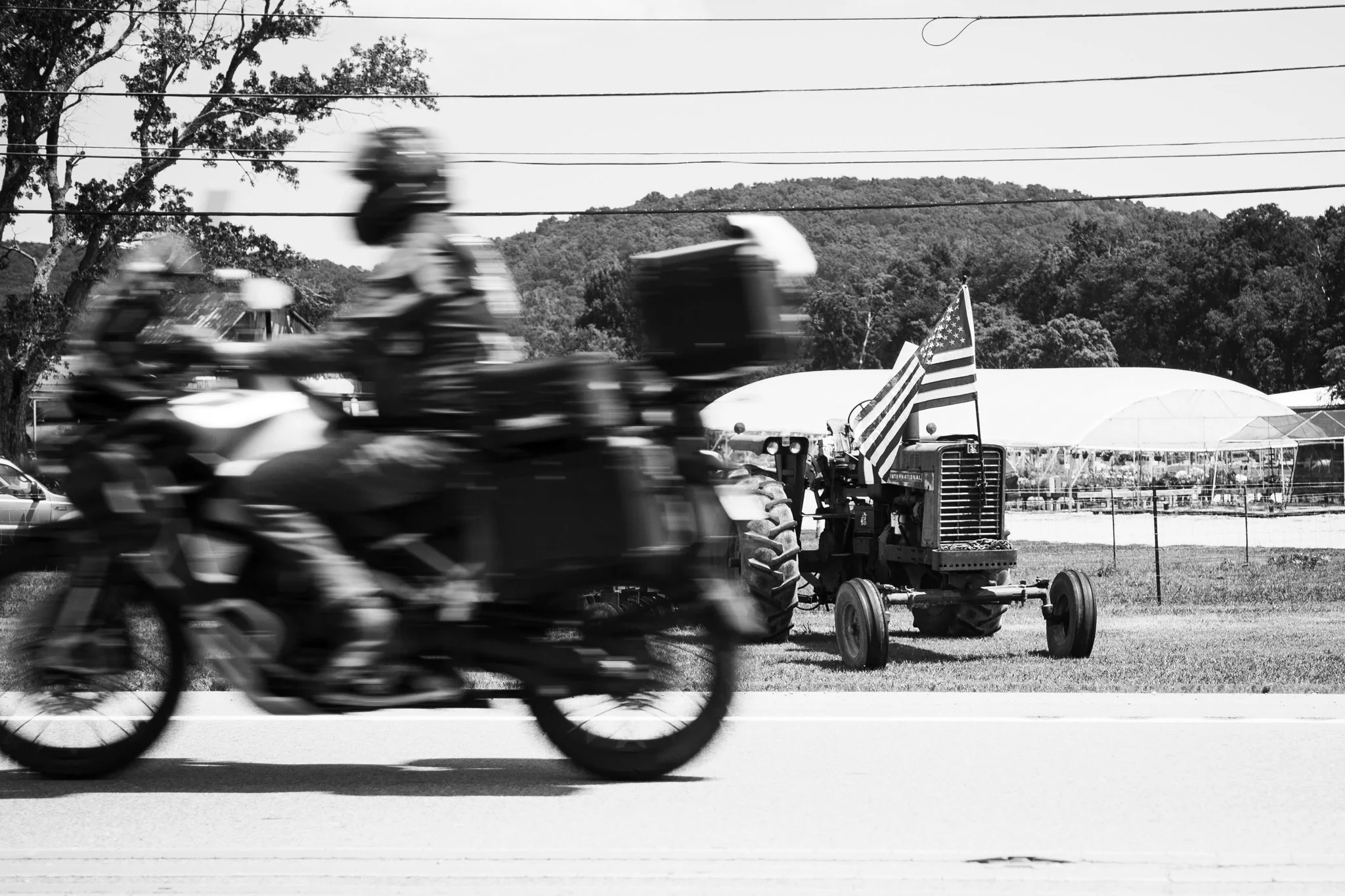 Black and white image of a motorcycle with a blurred rider speeding along a road, with a tractor and flags visible in the background.