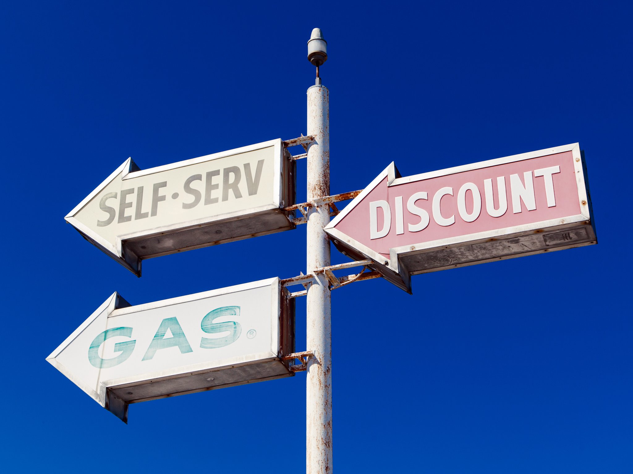 Old gas station sign with arrows indicating self-service and discount, mounted on a rusted pole against a clear blue sky.