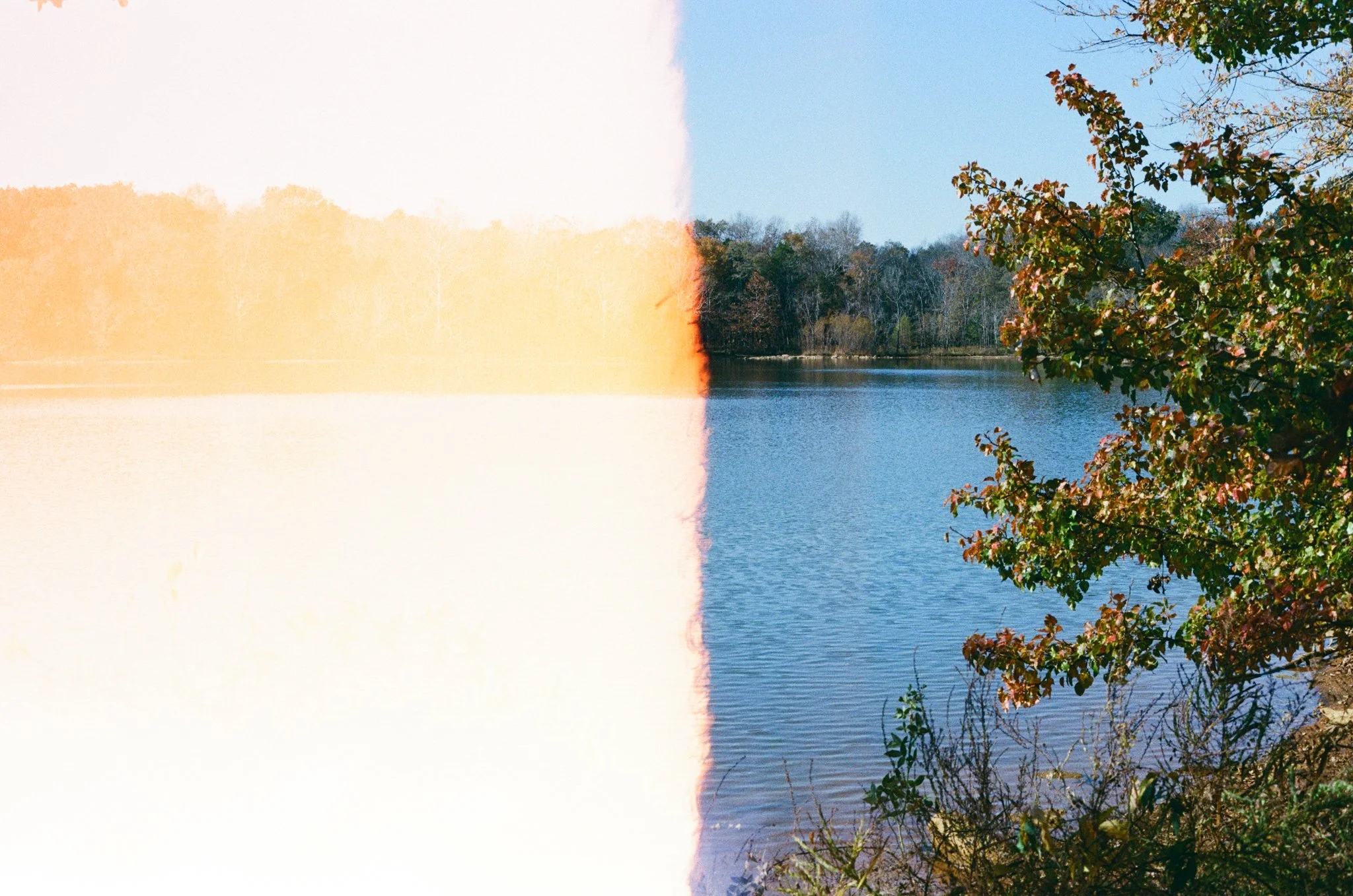 A serene lakeside scene with calm water, trees with autumn foliage, and a partly cloudy blue sky, with one side of the image overexposed and faded.