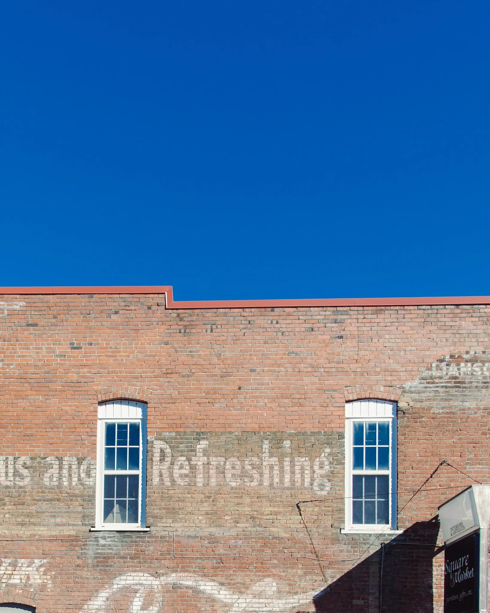 Brick building with three tall windows on the upper floor, a brick facade with some weathering, a sign for a market or antique store, and a clear blue sky.