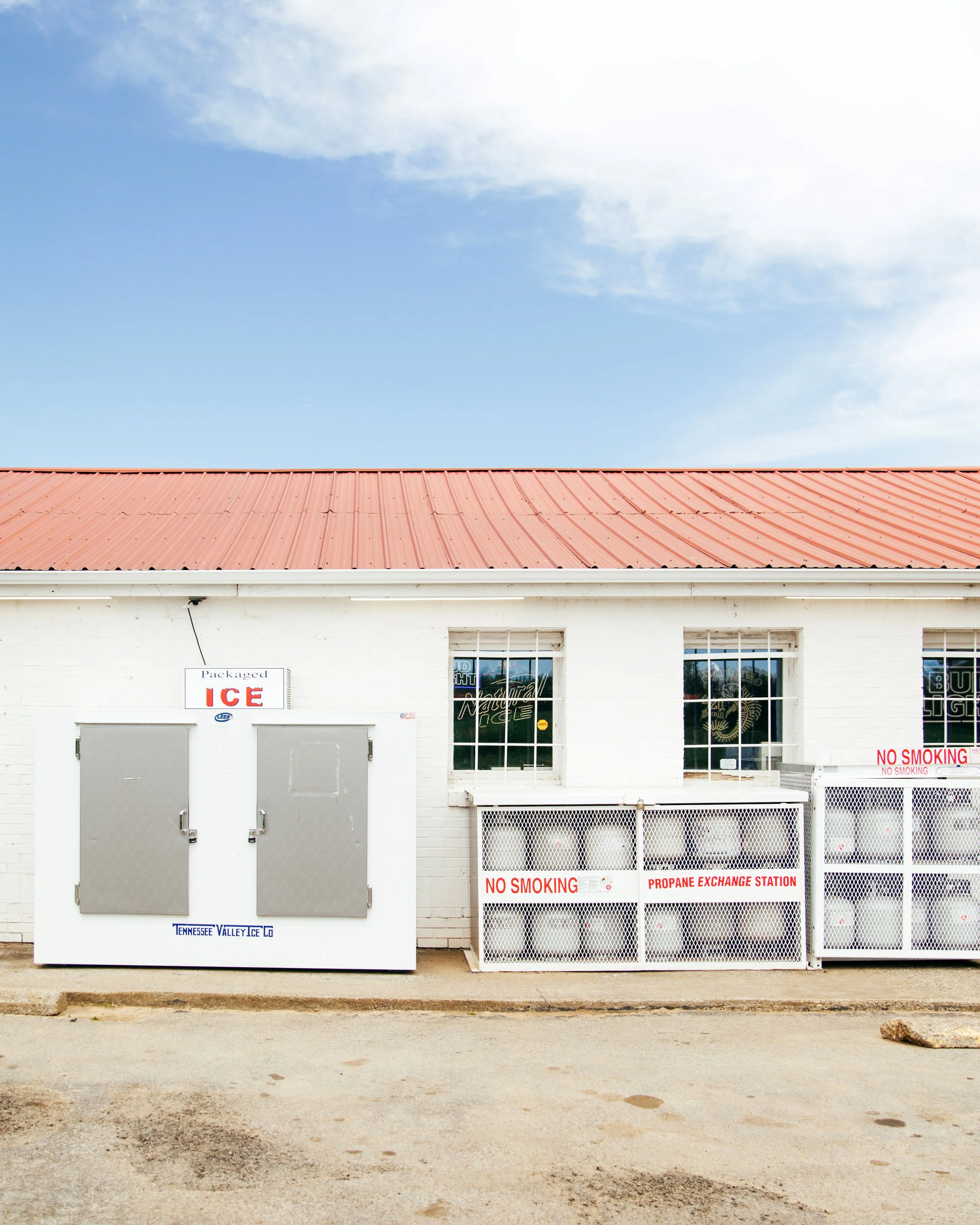 A white building with a red metal roof, two large gray metal doors, a window, and signs indicating it is a propane exchange station, including a 'No Smoking' sign and a propane exchange sign. There is a gravel ground in front of the building and a cl