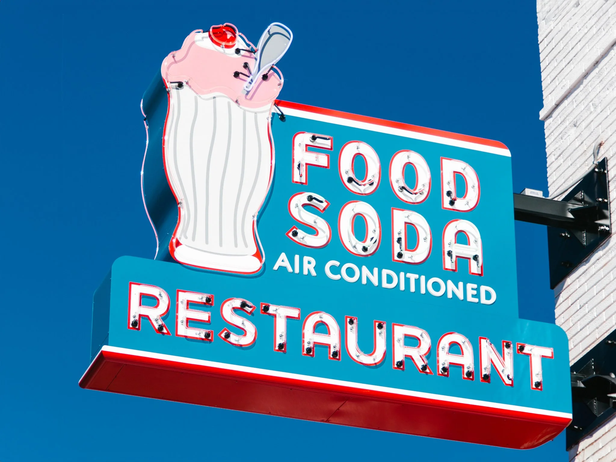 Colorful neon sign for a restaurant, featuring a milkshake with a cherry on top and the words 'Food Stop' and 'Restaurante', mounted on a building against a clear blue sky.