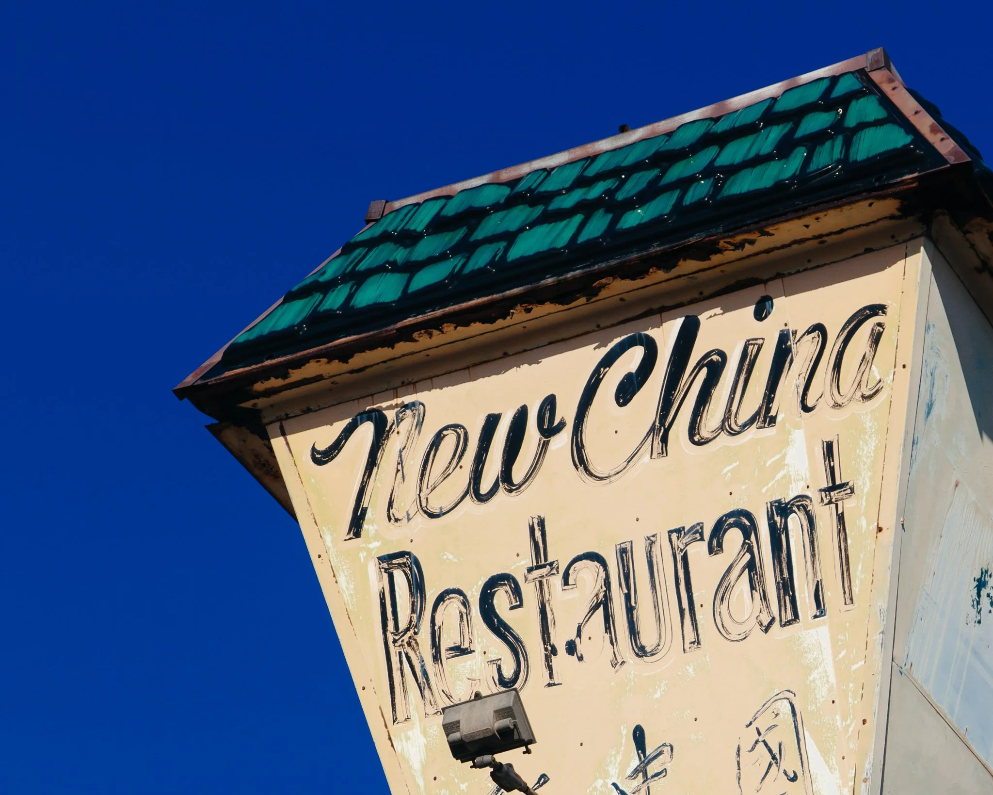 Close-up of an old, weathered sign with partially visible handwritten text against a clear blue sky.