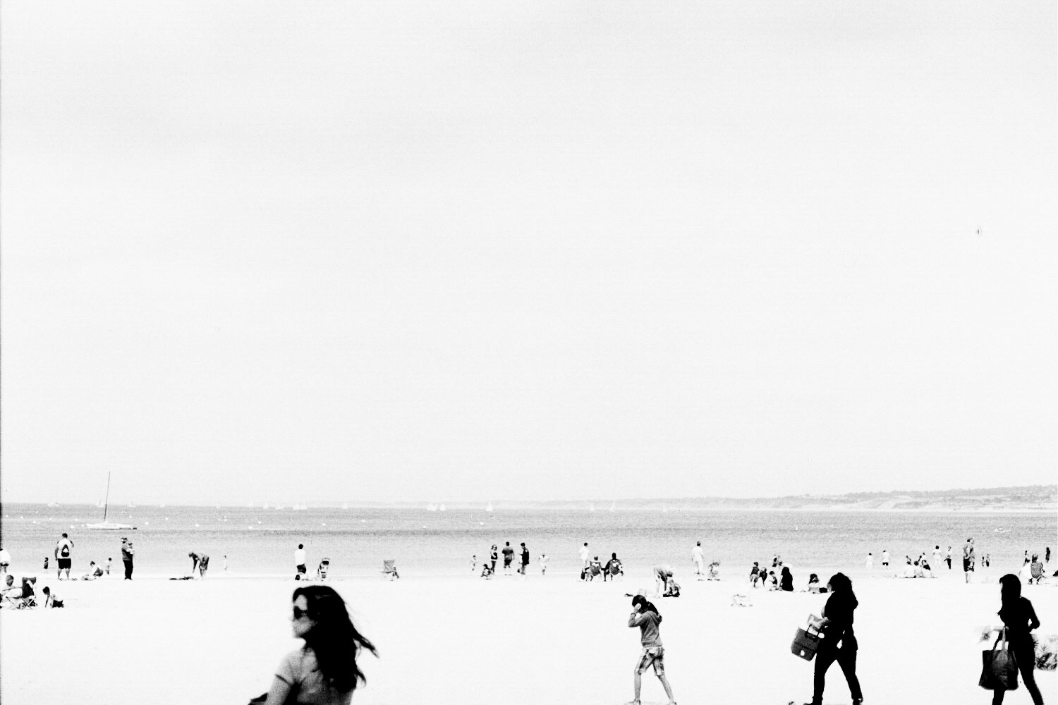 Black-and-white photo of people at the beach, some sitting, some walking, with the ocean in the background and a sailboat on the water.
