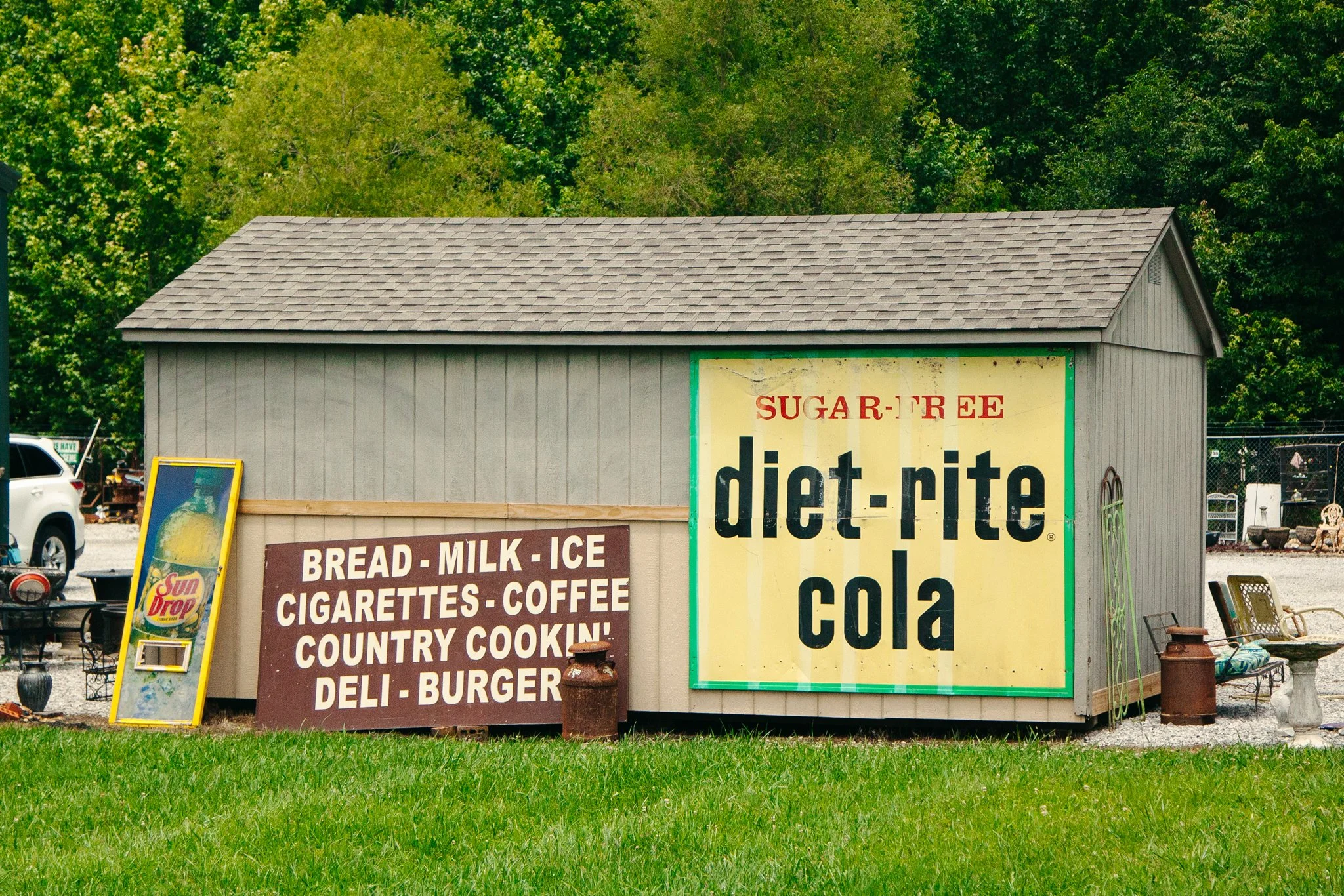 A small building with signs advertising baked goods, beverages, and country cooking, including bread, milk, ice, cigarettes, coffee, and burgers, with lush green trees in the background.