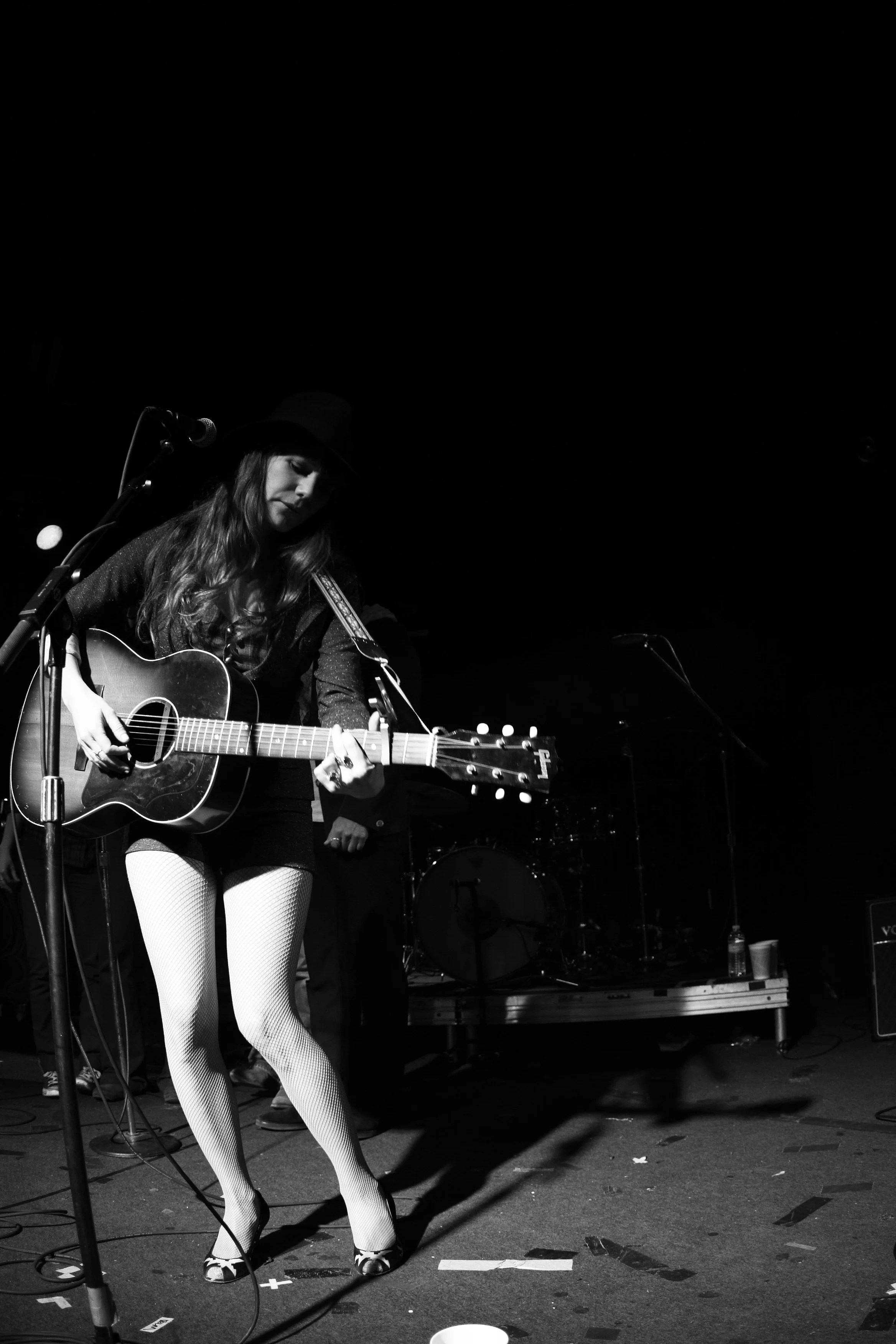 A woman playing an acoustic guitar on stage, wearing a hat, white fishnet tights, and patterned shoes, with a microphone in front of her.