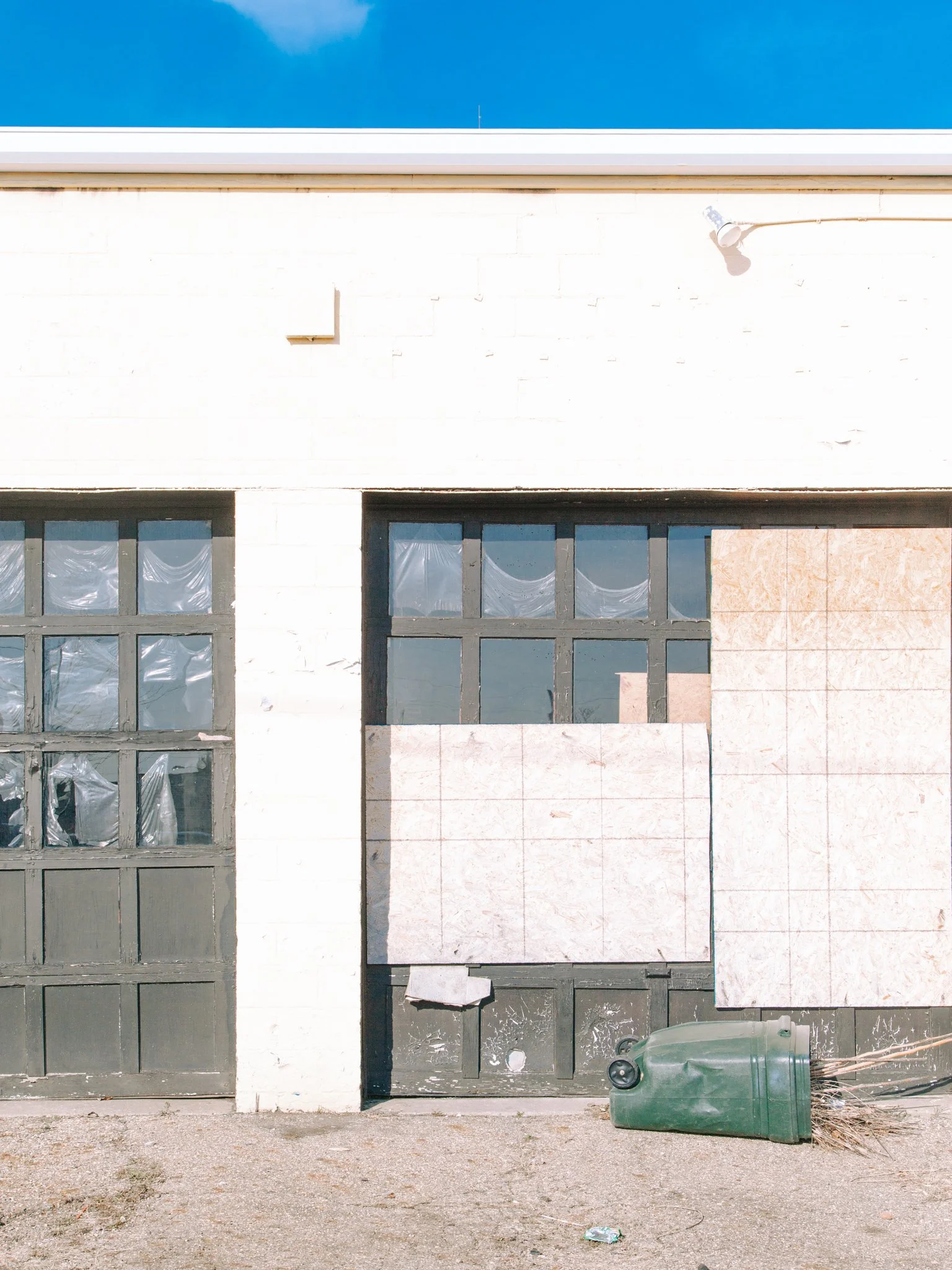 A building with a facade of white bricks and large black windowpanes, a door, and a white division. A green trash bin with arms and tied to a small bush stands in front of the building, on a gravel or dirt pathway with some debris on the ground.