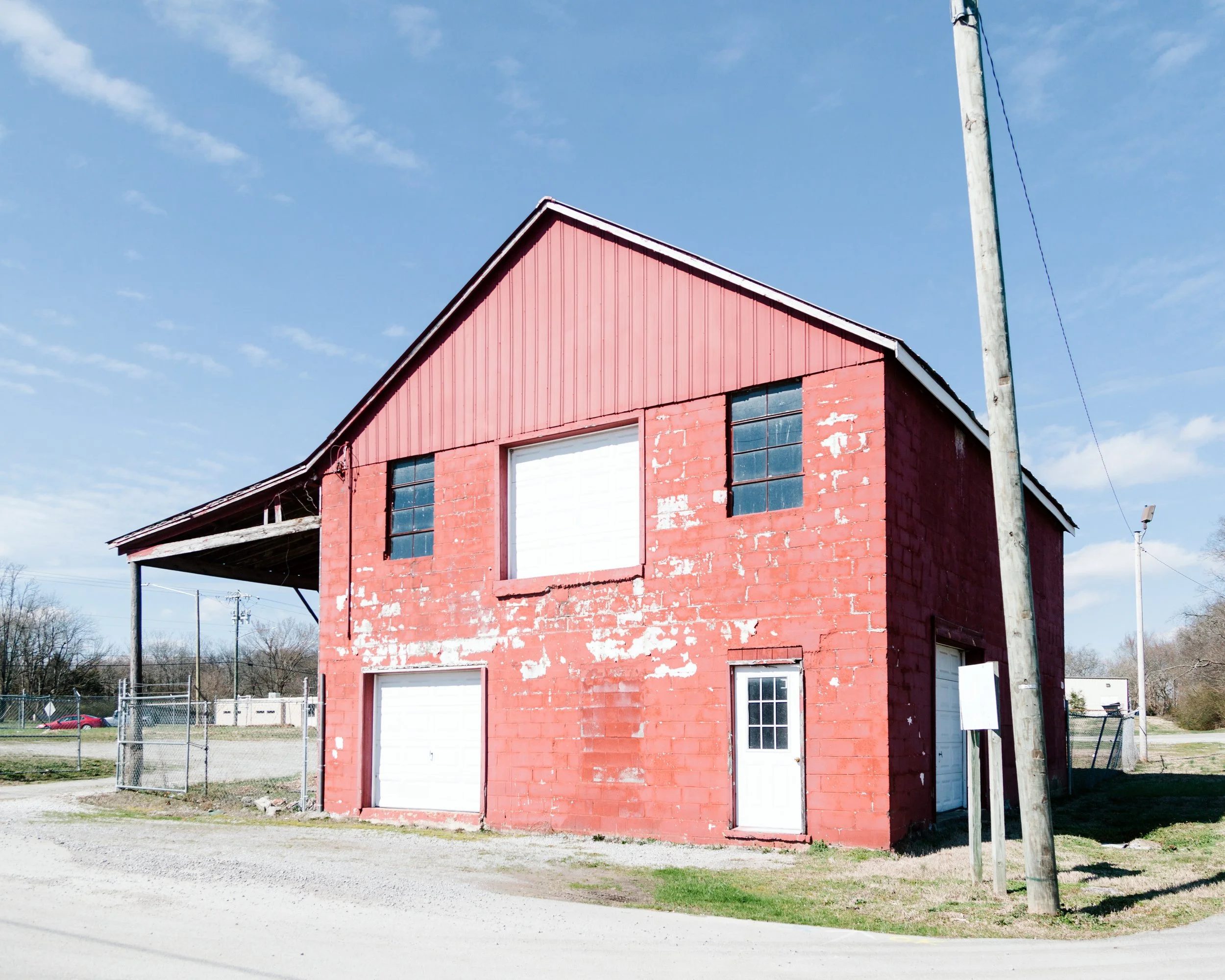 A red brick building with white garage doors and windows, fronted by a gravel area and a fenced yard, with utility poles and power lines against a clear blue sky.