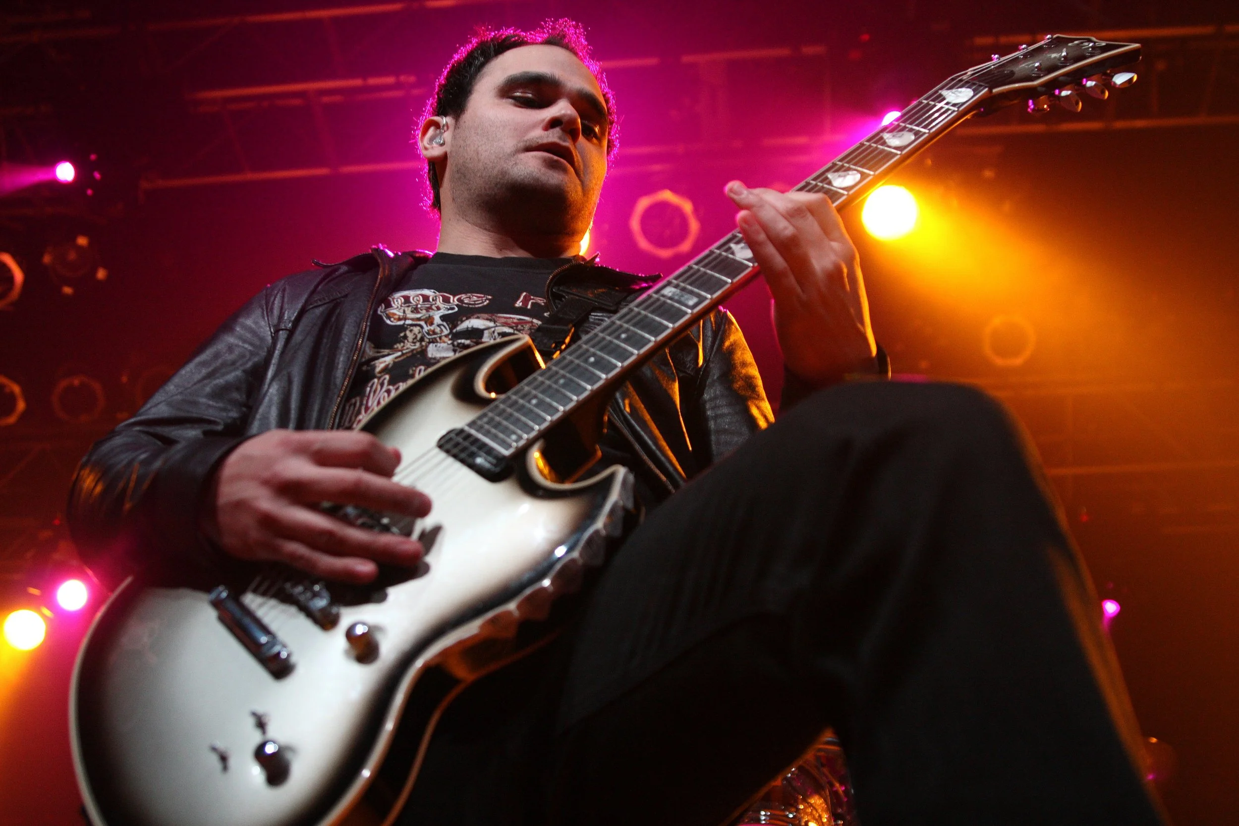 A male guitarist in a leather jacket performing on stage with colorful stage lighting in pink, yellow, and orange hues.
