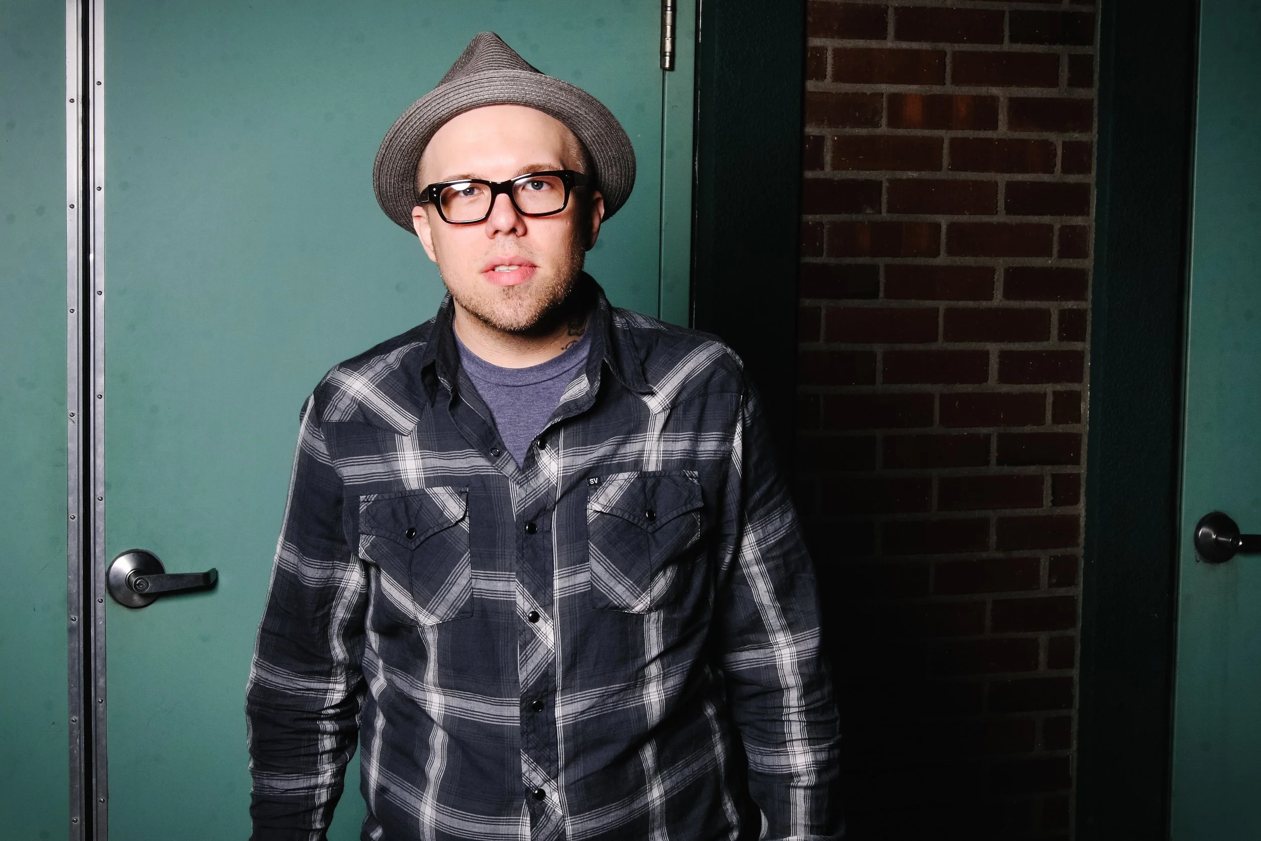 A man with glasses, a beard, and a mustache wearing a gray fedora hat and a plaid shirt standing in front of a green door and brick wall.