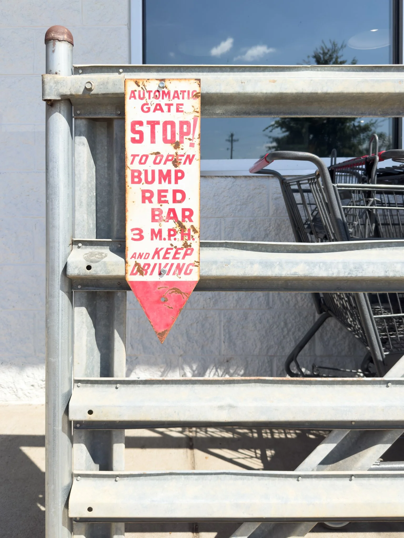 A weathered metal sign on a shopping cart corral that reads 'Automatic Gate Stop! To Open Bump Red Bar 3 M.P.H. and Keep Driving' in red text, with shopping carts visible behind the sign and a building with a window in the background.