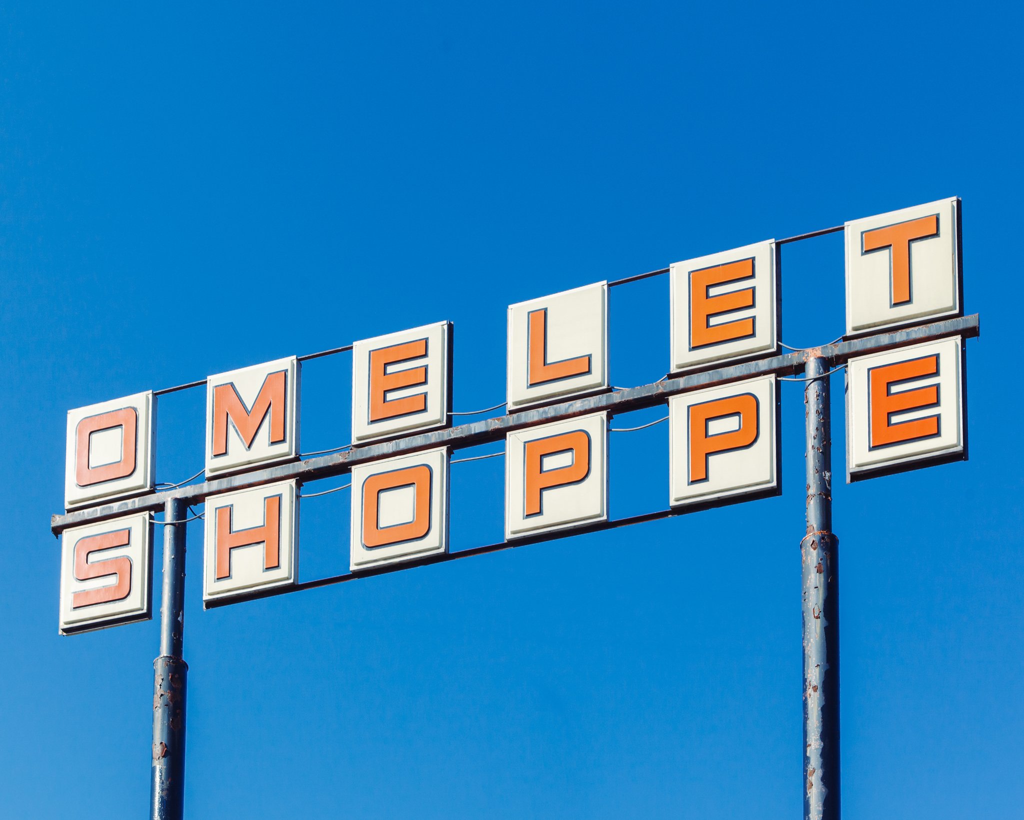 A vintage roadside motel sign with orange letters on white background, mounted on metal poles against a clear blue sky.