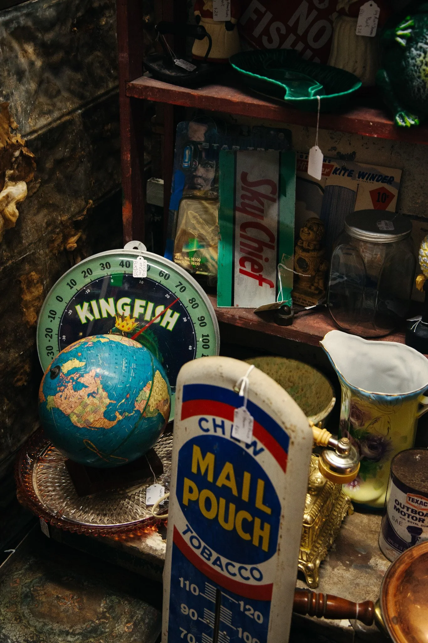 A shelf with various objects including a globe, a clock labeled 'KINGFISH,' a tin with a picture of a man, some bags, a jar, a brass lamp, and a thermometer with a 'MAIL POUCH TOBACCO' sign hanging in the foreground.