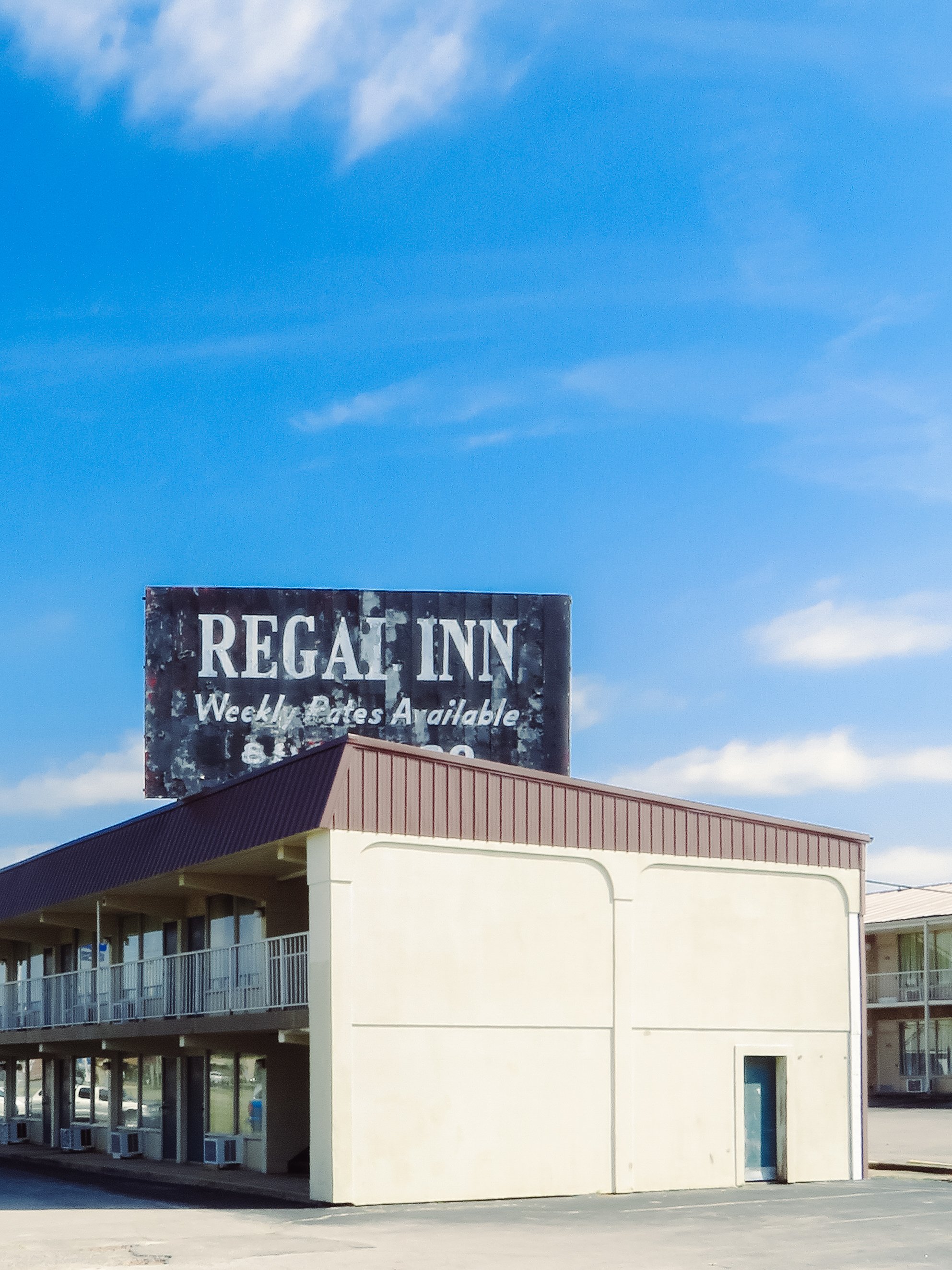 A two-story motel with exterior corridors and a beige facade, featuring a visible sign for Regal Inn at the top, against a blue sky with some clouds.