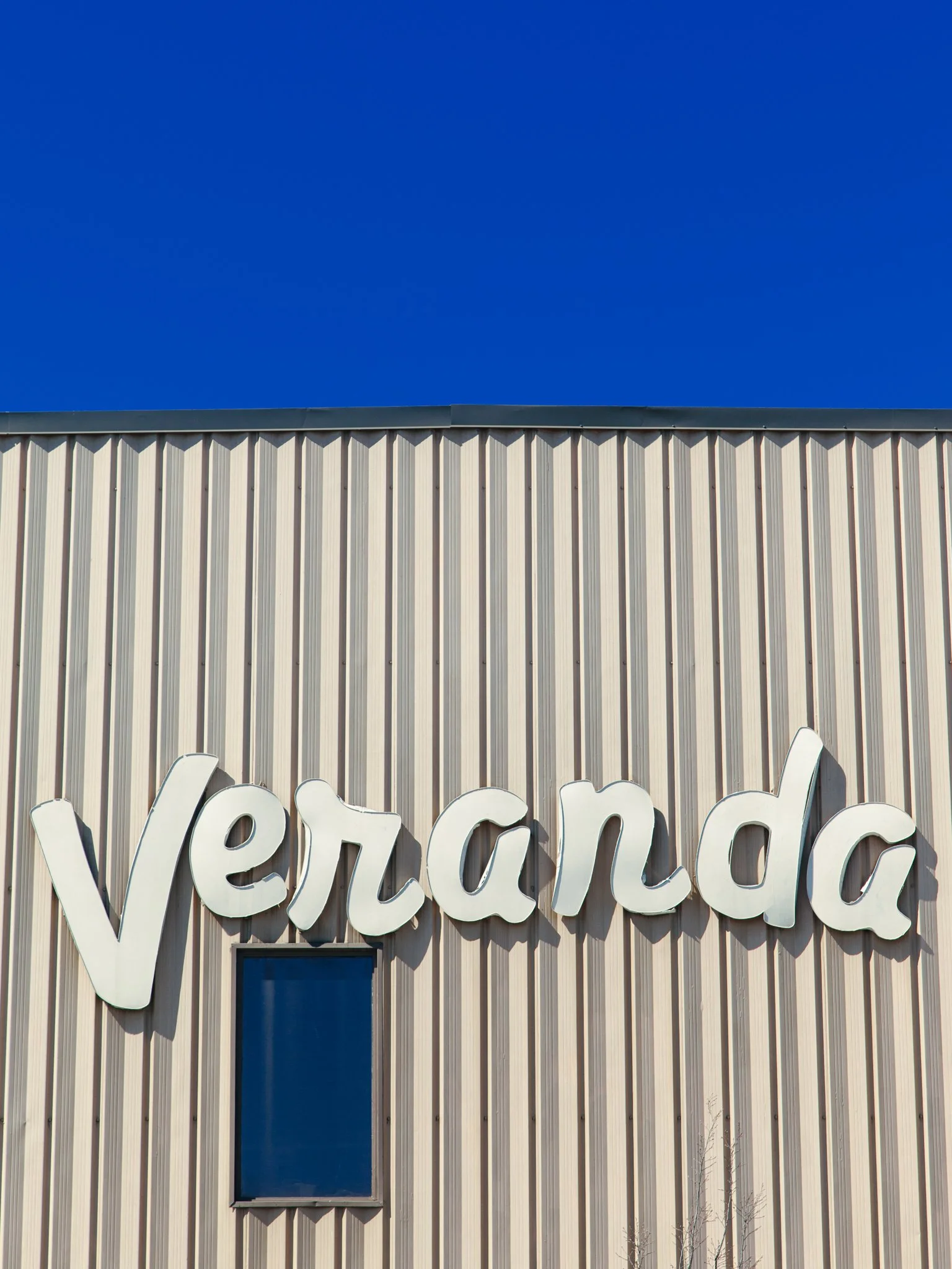 Part of a beige building with vertical metal siding, a black-framed window, and large white letters spelling out "Vero" on the facade under a clear blue sky.