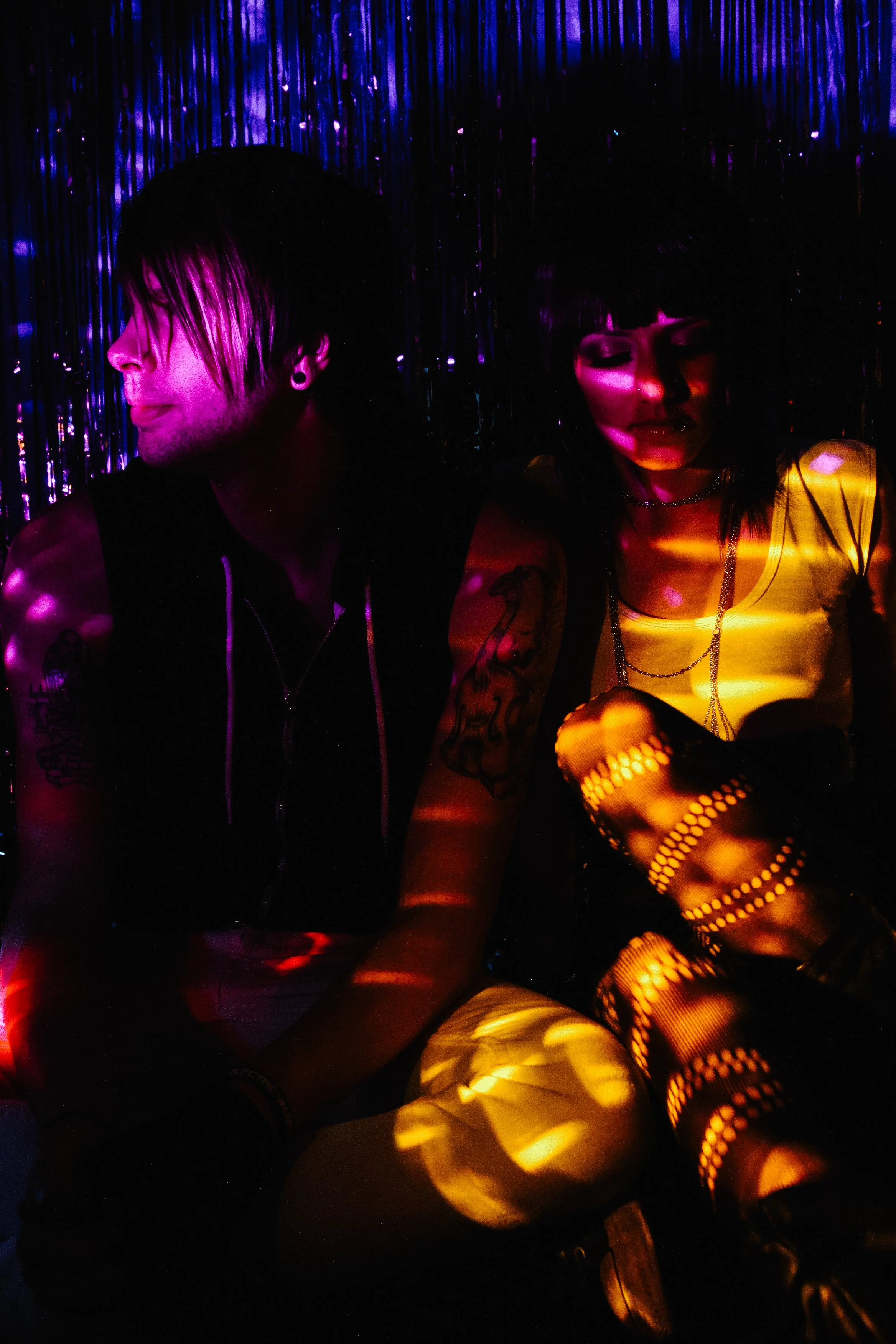 Two people sitting against a background of shiny purple and blue streamers, illuminated by colorful, dotted lighting.