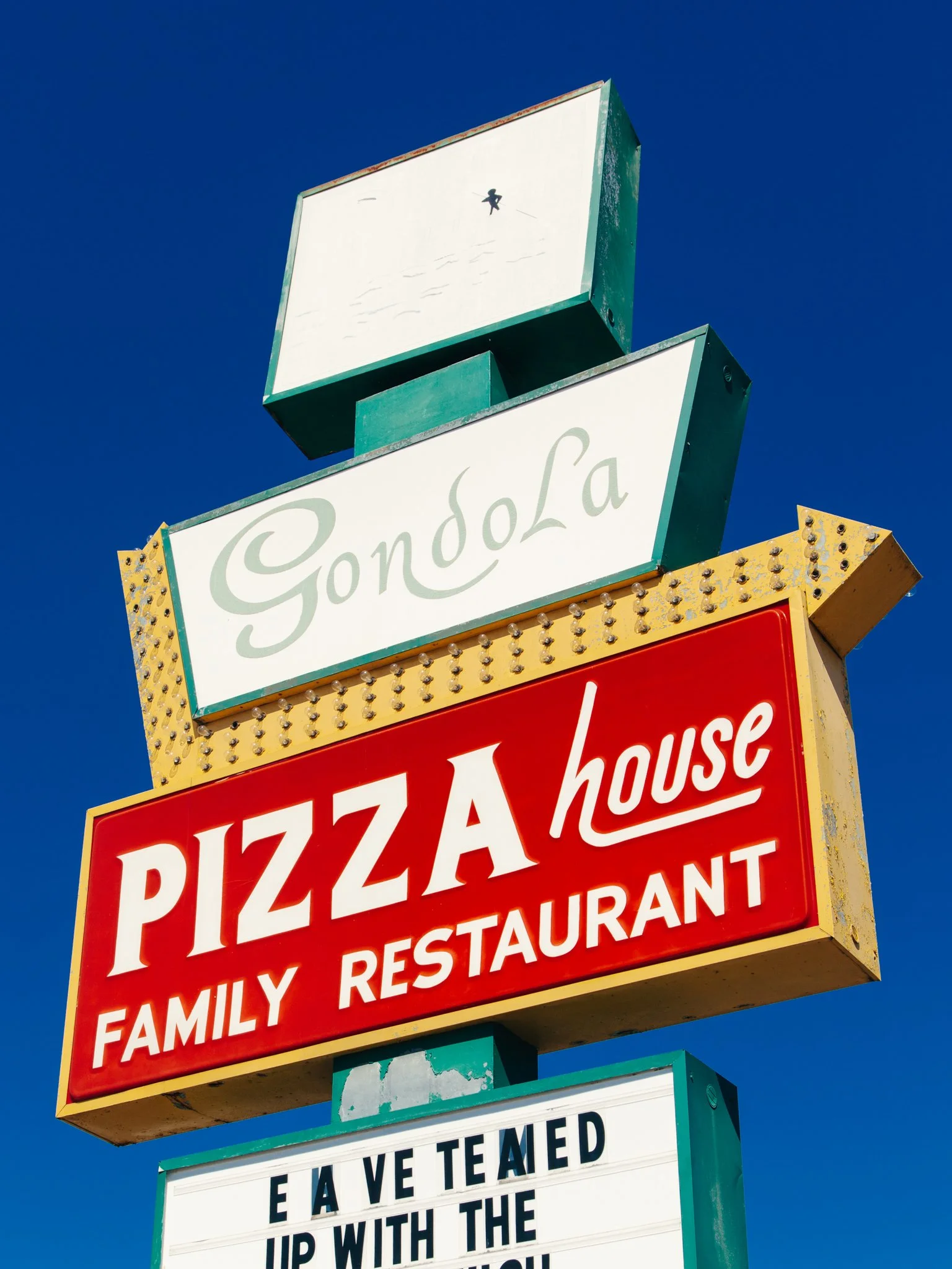 A vintage-style sign for a family restaurant and pizzeria called Gondola, with a small sign above displaying a doll figure and a white sign below with black text, against a clear blue sky.