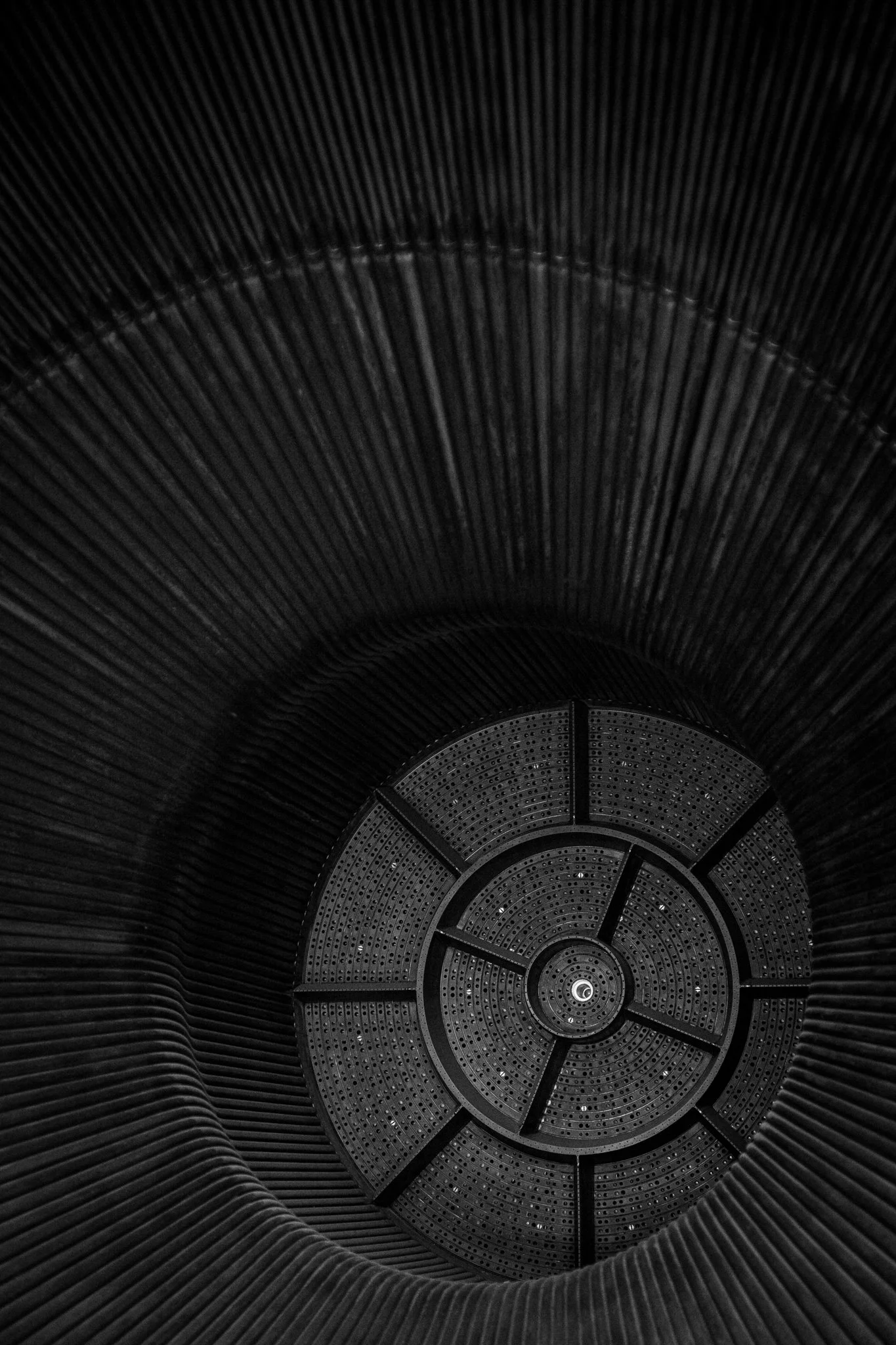 Looking down into a modern architectural tunnel with concentric circular design, dark metallic interior, and detailed patterns on the floor.