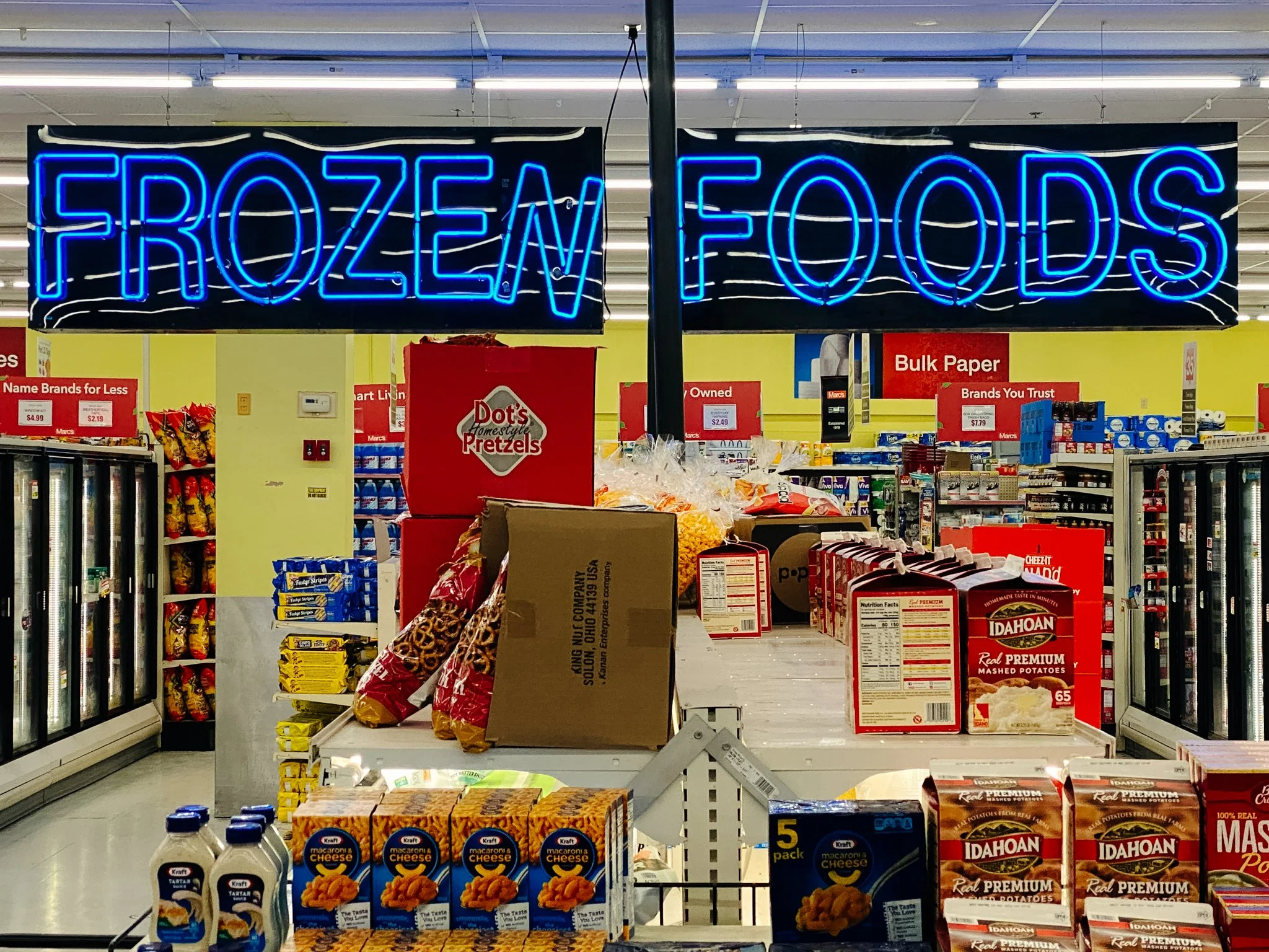 Neon sign with blue letters displaying 'FROZEN FOOD' hanging above grocery store aisles filled with various packaged food items and snacks.