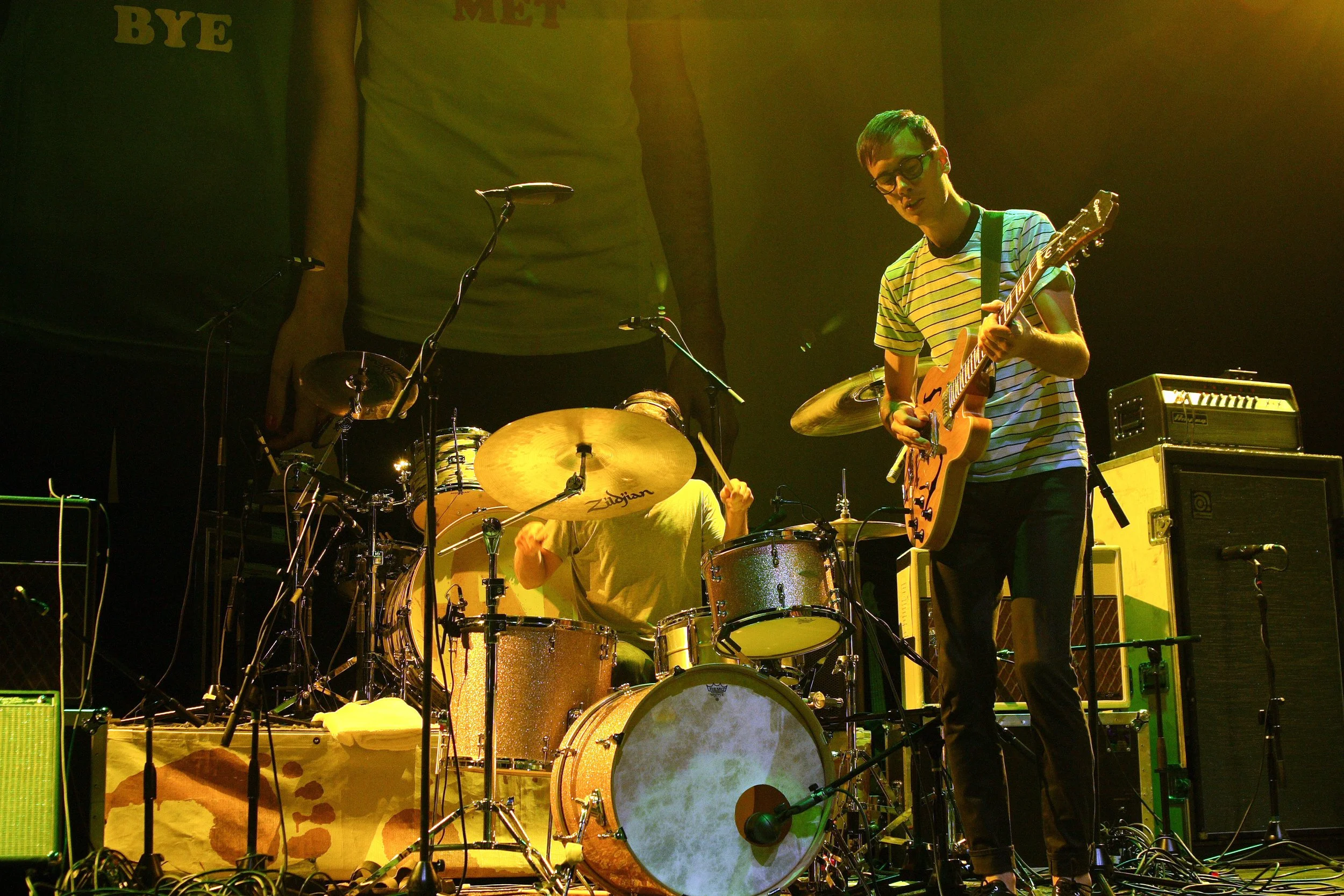 Musicians performing on stage with drums, guitar, and amplifiers, in a dimly lit setting.