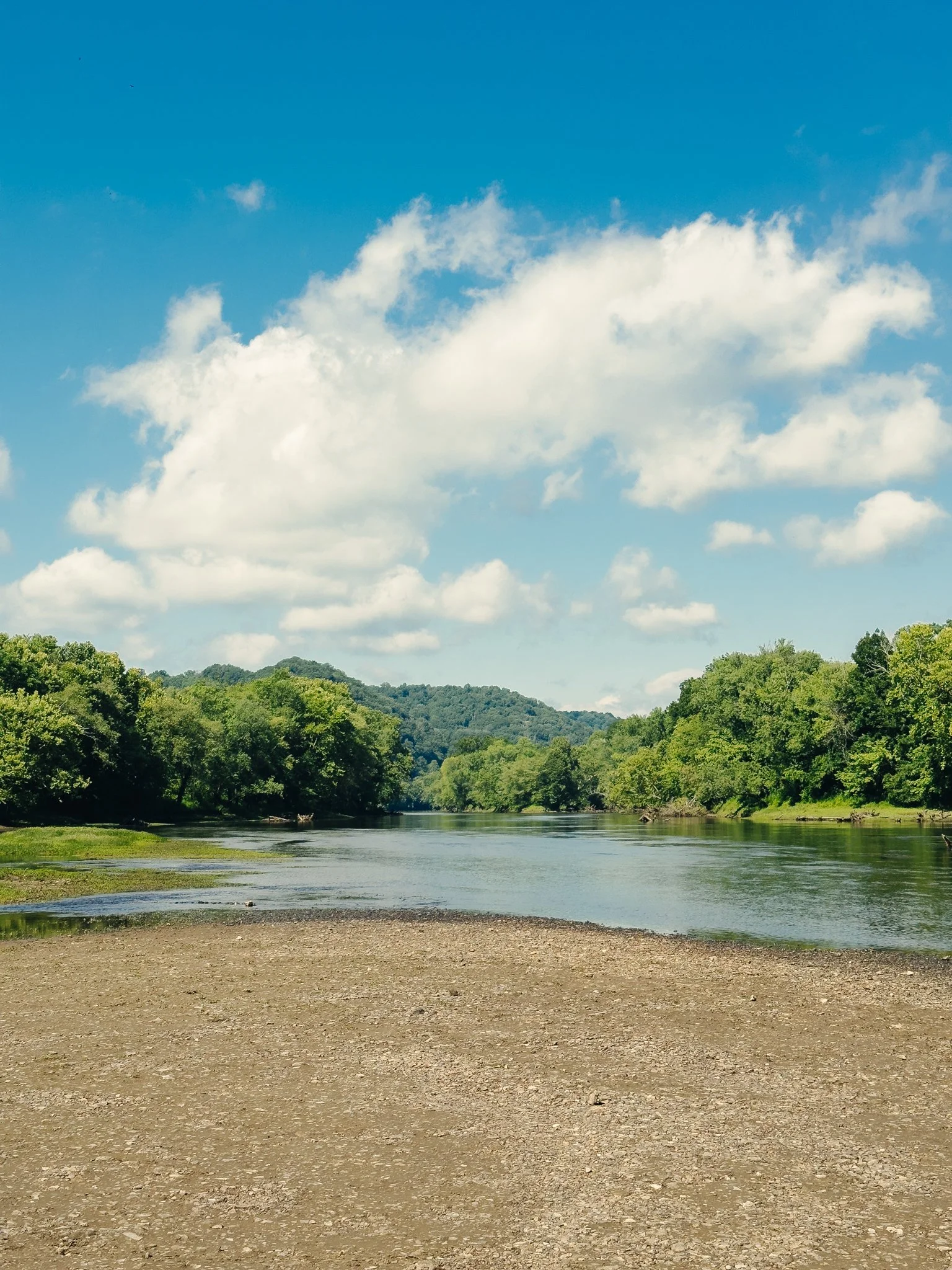 A peaceful river scene with calm water surrounded by green trees and hills under a blue sky with white clouds.