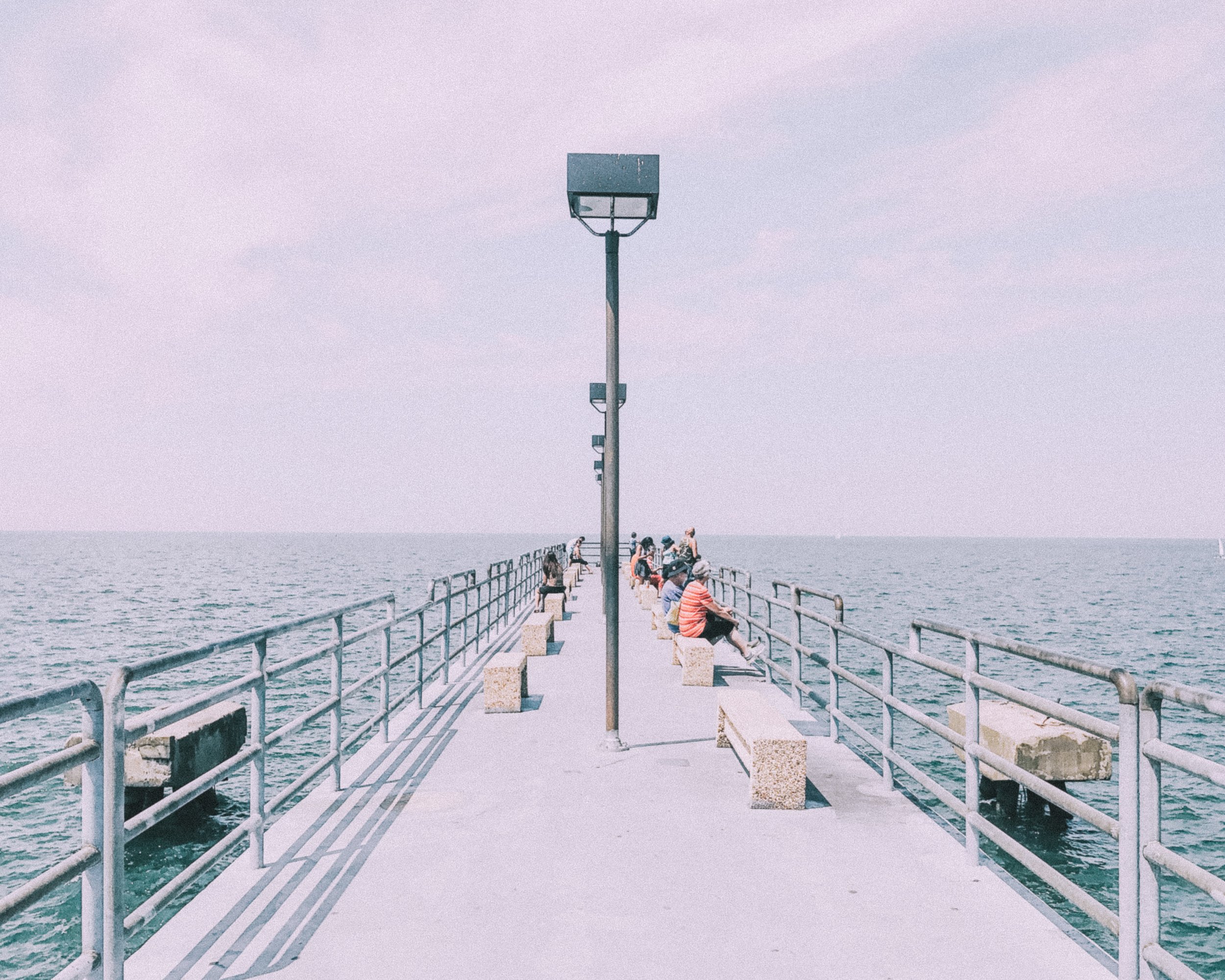 A group of people sitting on benches and standing on a pier extending into a large body of water, with a sky above and a tall lamppost in the center.