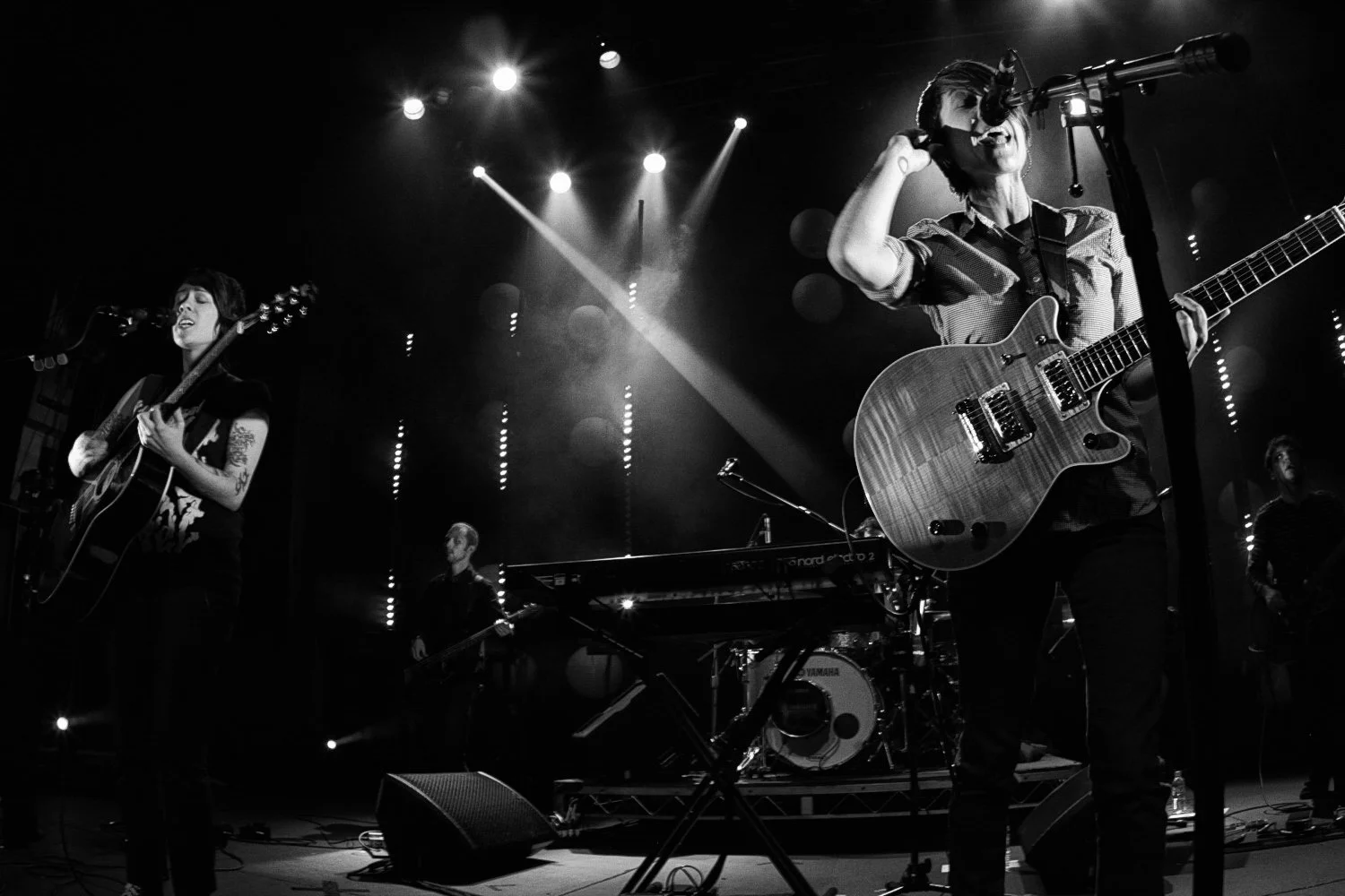 Black and white photo of a band performing live on stage with multiple musicians playing guitars, keyboard, and drums under stage lighting.