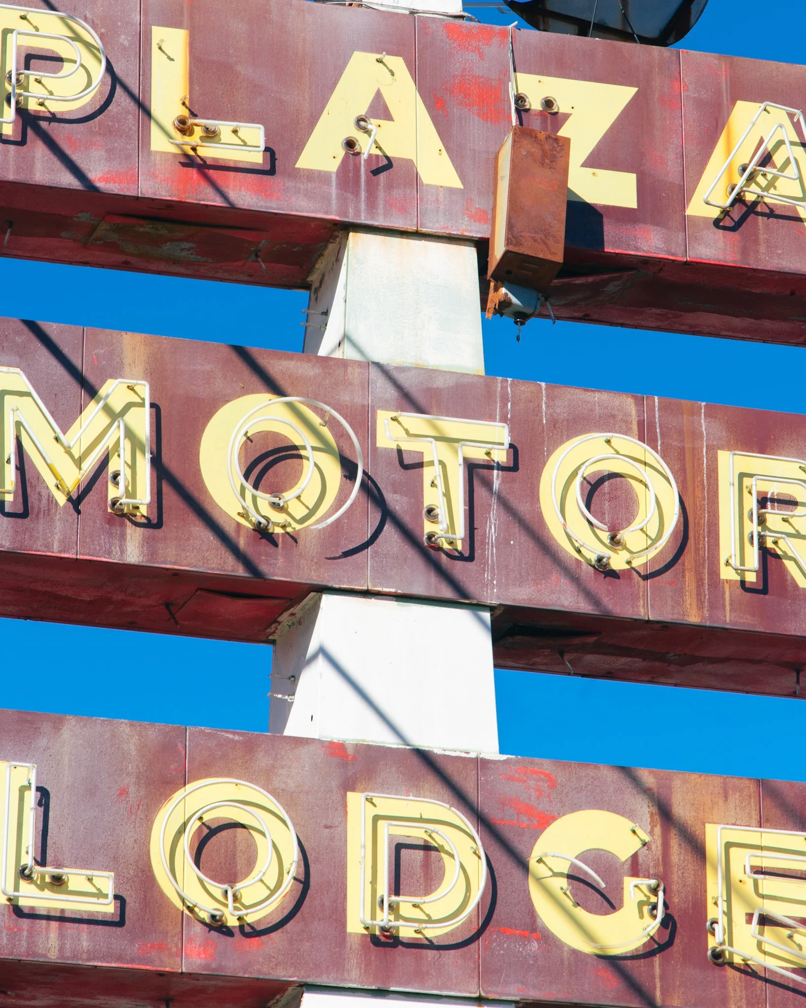 Close-up of vintage neon sign with partially visible yellow lettering on a red background, mounted on a white pole against clear blue sky.