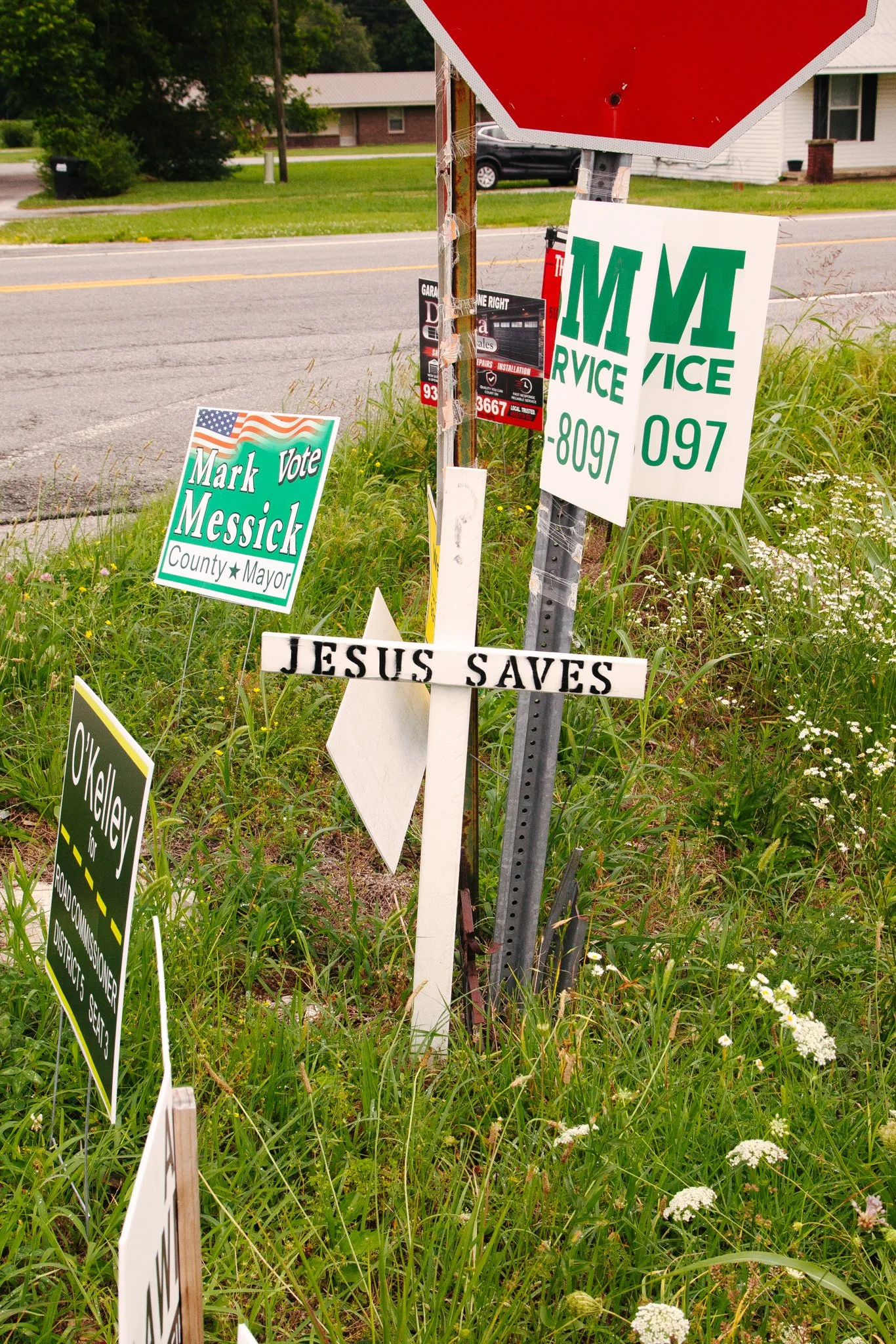 Multiple campaign signs in a grassy area beside a road, including a green sign for Mark Messick, a white and green sign with the abbreviation 'M M' and the word 'Service,' and a white cross-shaped sign that reads 'Jesus Saves.'