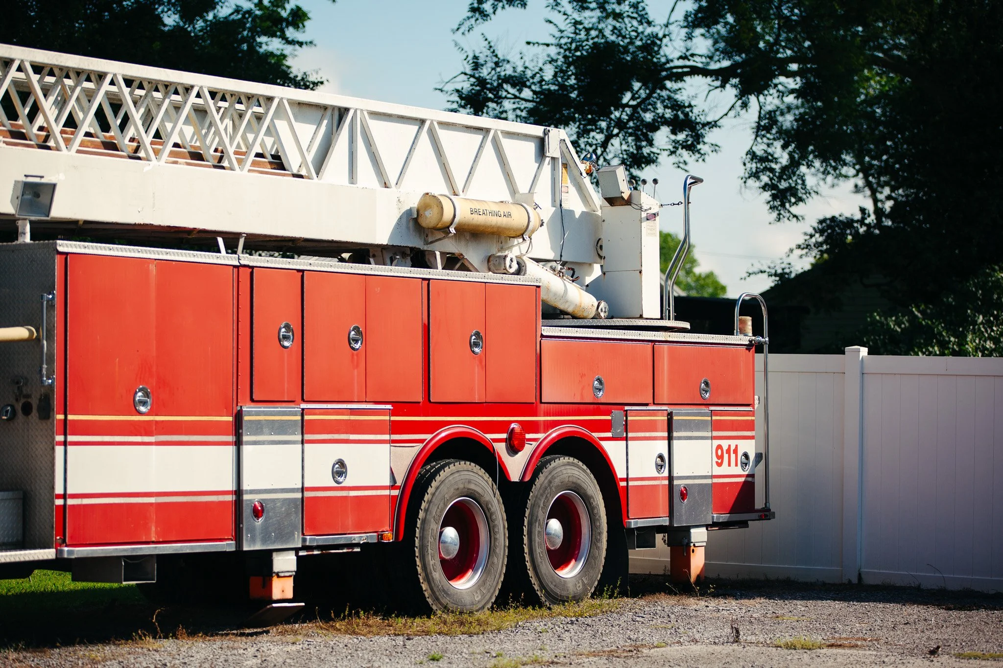 A fire truck with an extended ladder on top, parked outdoors in front of a white fence and trees.