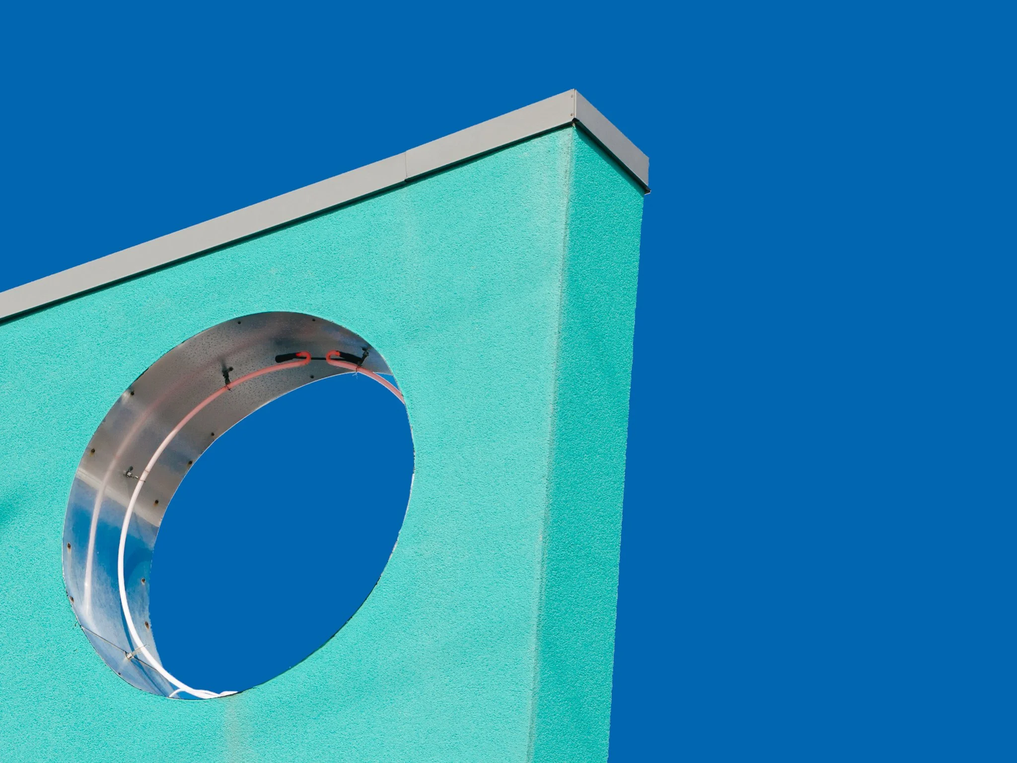 Close-up of a modern architectural structure with a circular opening, mounted on a textured turquoise wall against a clear blue sky.