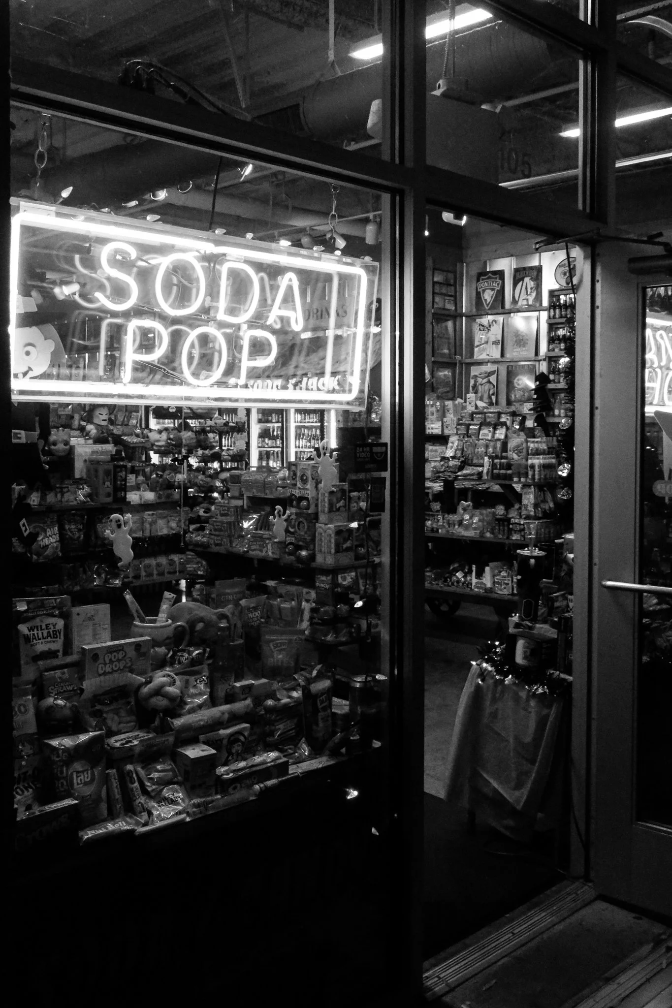 A view into a convenience store or small shop with a neon sign that reads 'SODA POP' in the window. The store displays various toys, snacks, and beverages, visible through the glass. The store's interior includes shelves stocked with items and poster