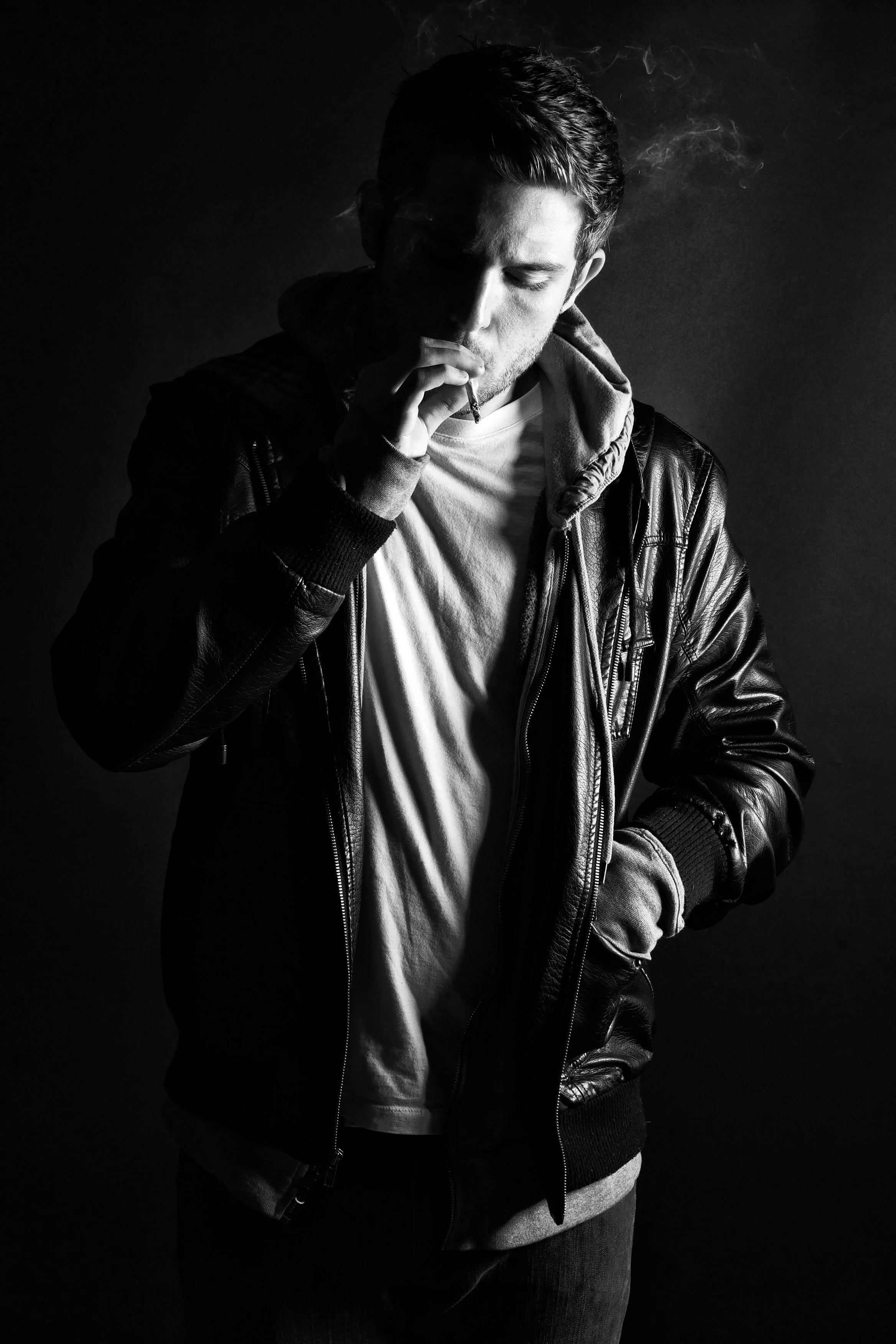 Black and white photo of a young man with short hair smoking a cigarette, wearing a leather jacket, white t-shirt, and dark pants, with a dark background.