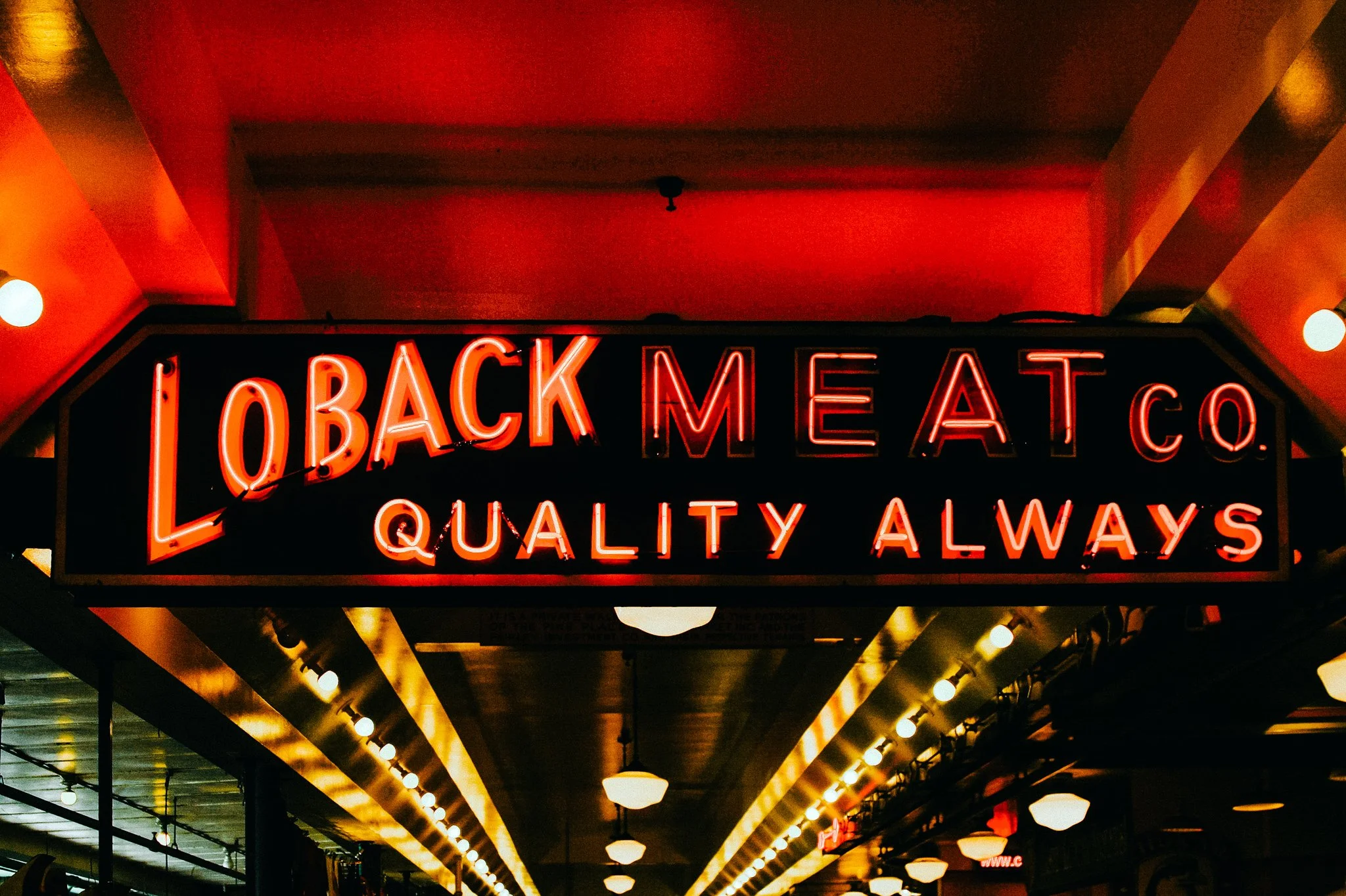 Neon sign reading "Loback Meat Co. Quality Always" hanging in a corridor with yellow lights and a ceiling with red lighting.