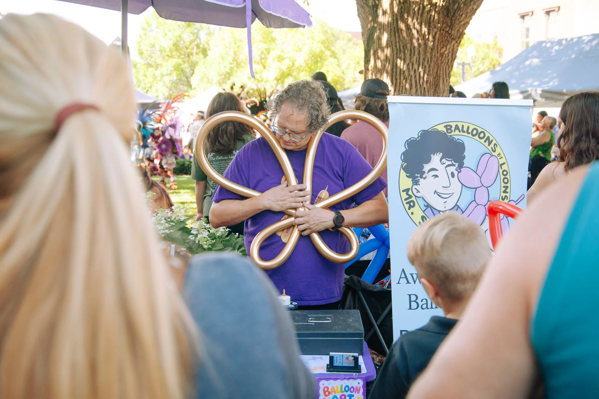 A man with gray curly hair and glasses making balloon art at an outdoor event, surrounded by children and adults. The man is wearing a purple shirt and holding golden balloon twists, with a colorful banner with a cartoon boy and balloons in the backg