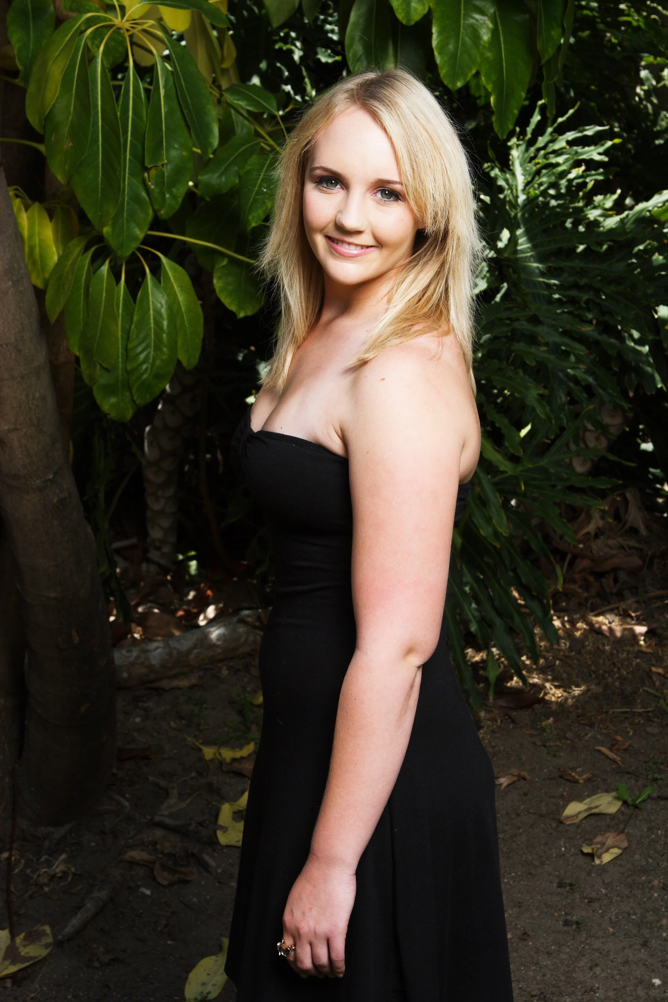 A young woman with blonde hair smiling, wearing a black strapless dress, standing outdoors among green tropical plants.