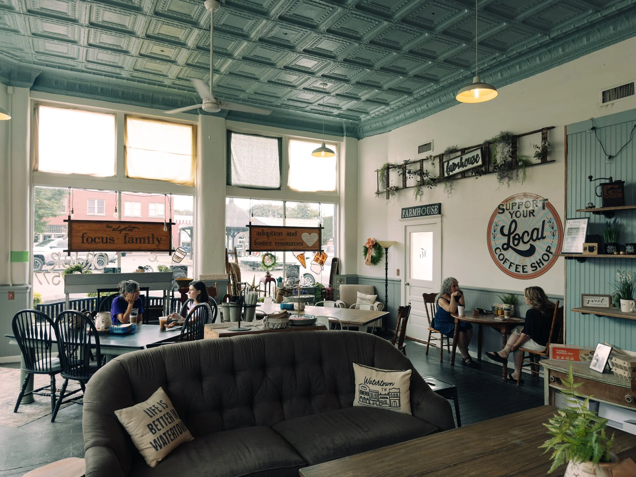 Interior of a cozy coffee shop with large windows, displaying hanging signs and plants, with customers seated and chatting.