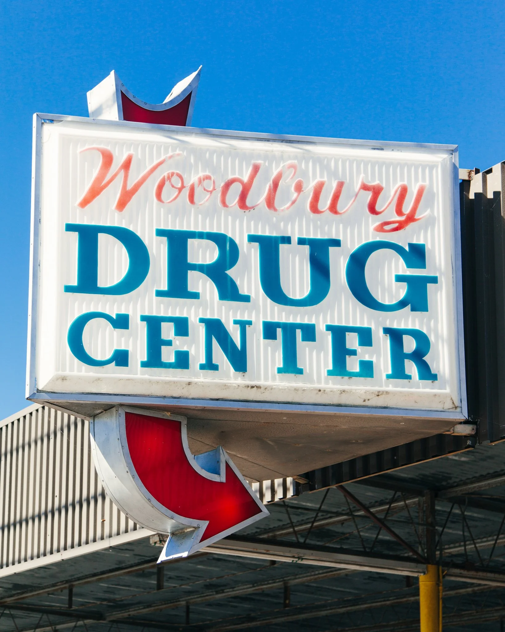 Neon sign reading 'Woodbury Drug Center' with a red and white arrow shape on a bright blue sky background.