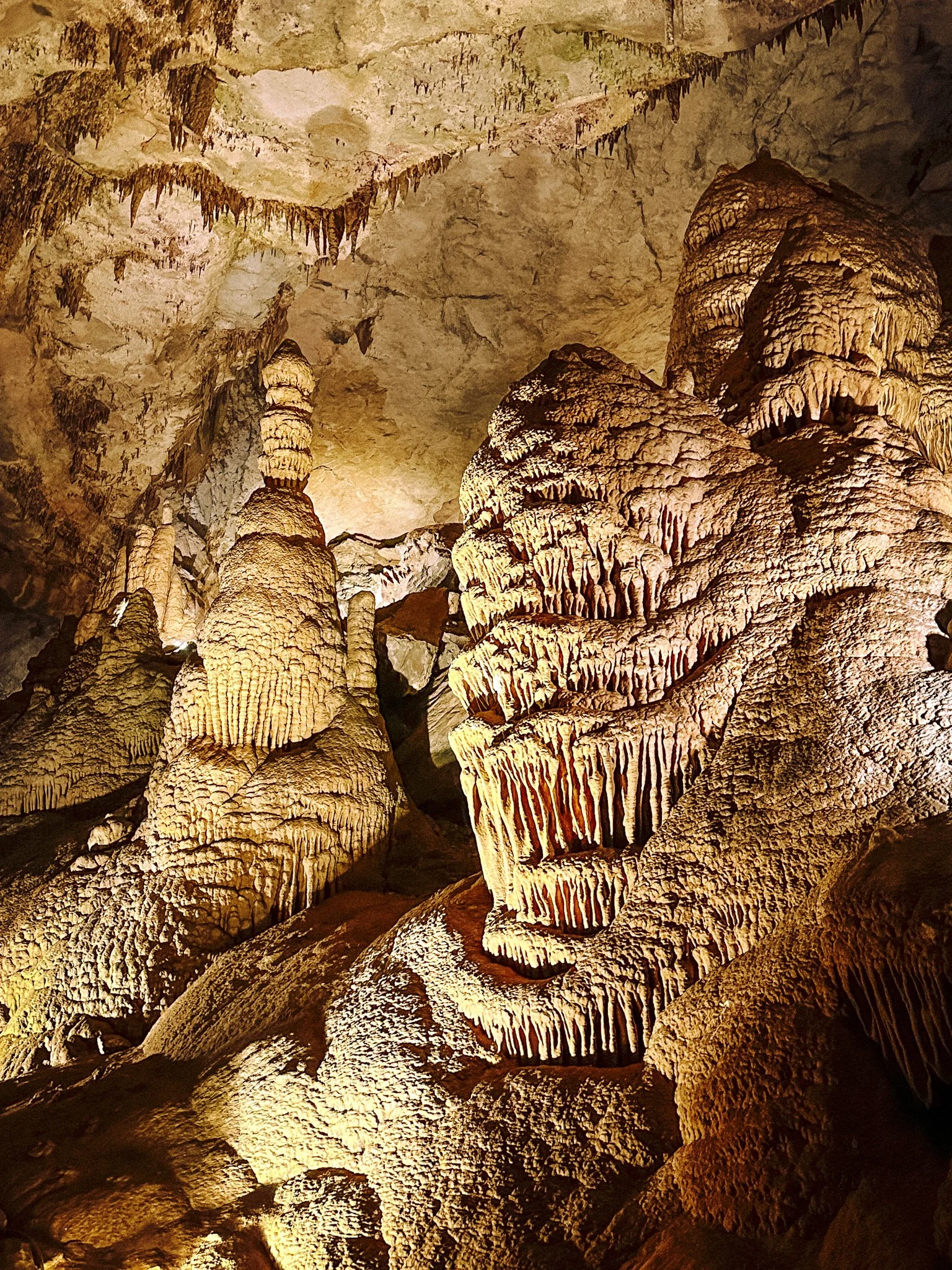 Inside view of a cave with stalactites hanging from the ceiling and stalagmites rising from the ground, illuminated by warm artificial lighting.
