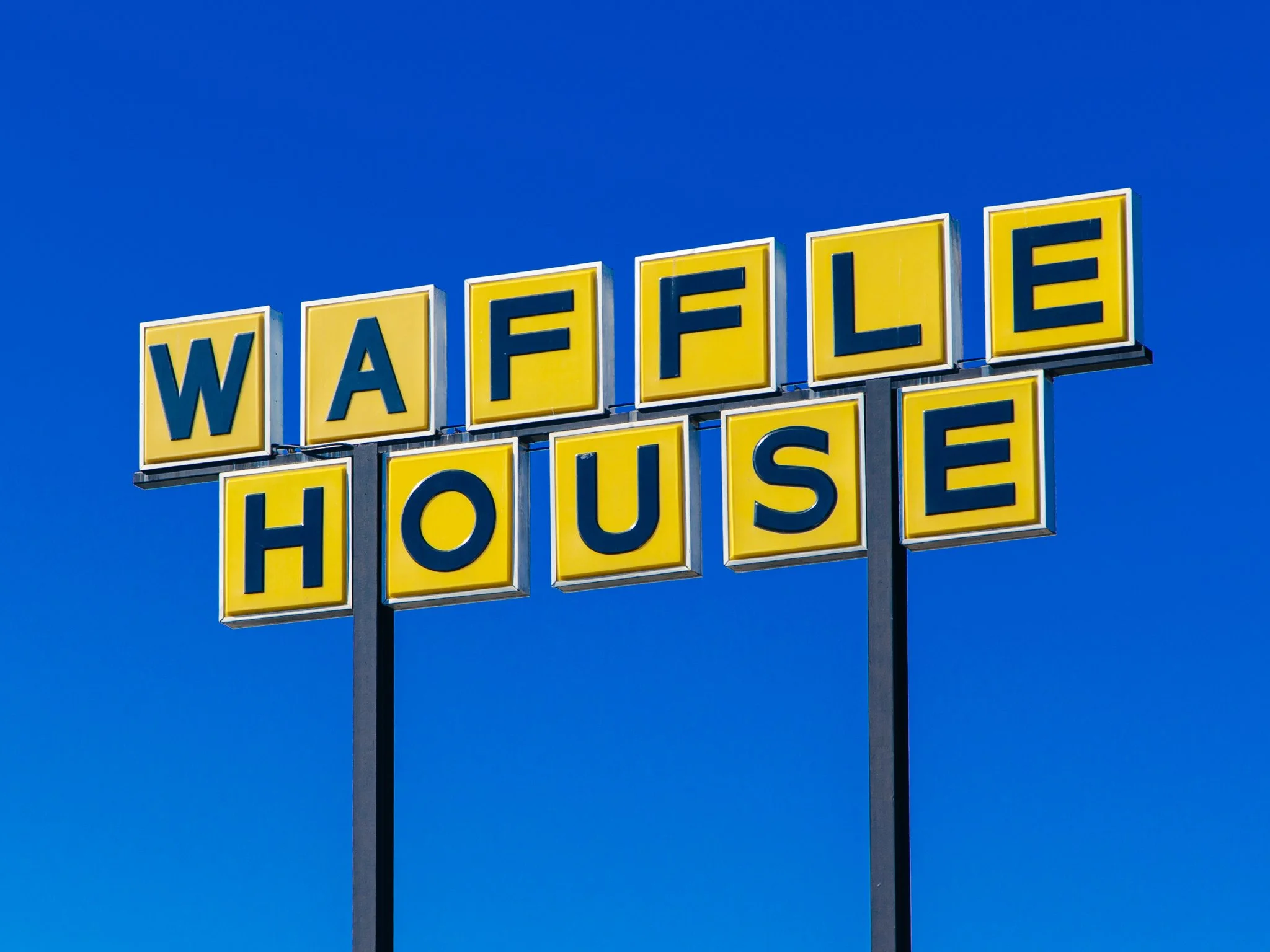 An outdoor sign with yellow background and blue letters spelling 'Waffle House' against a clear blue sky.