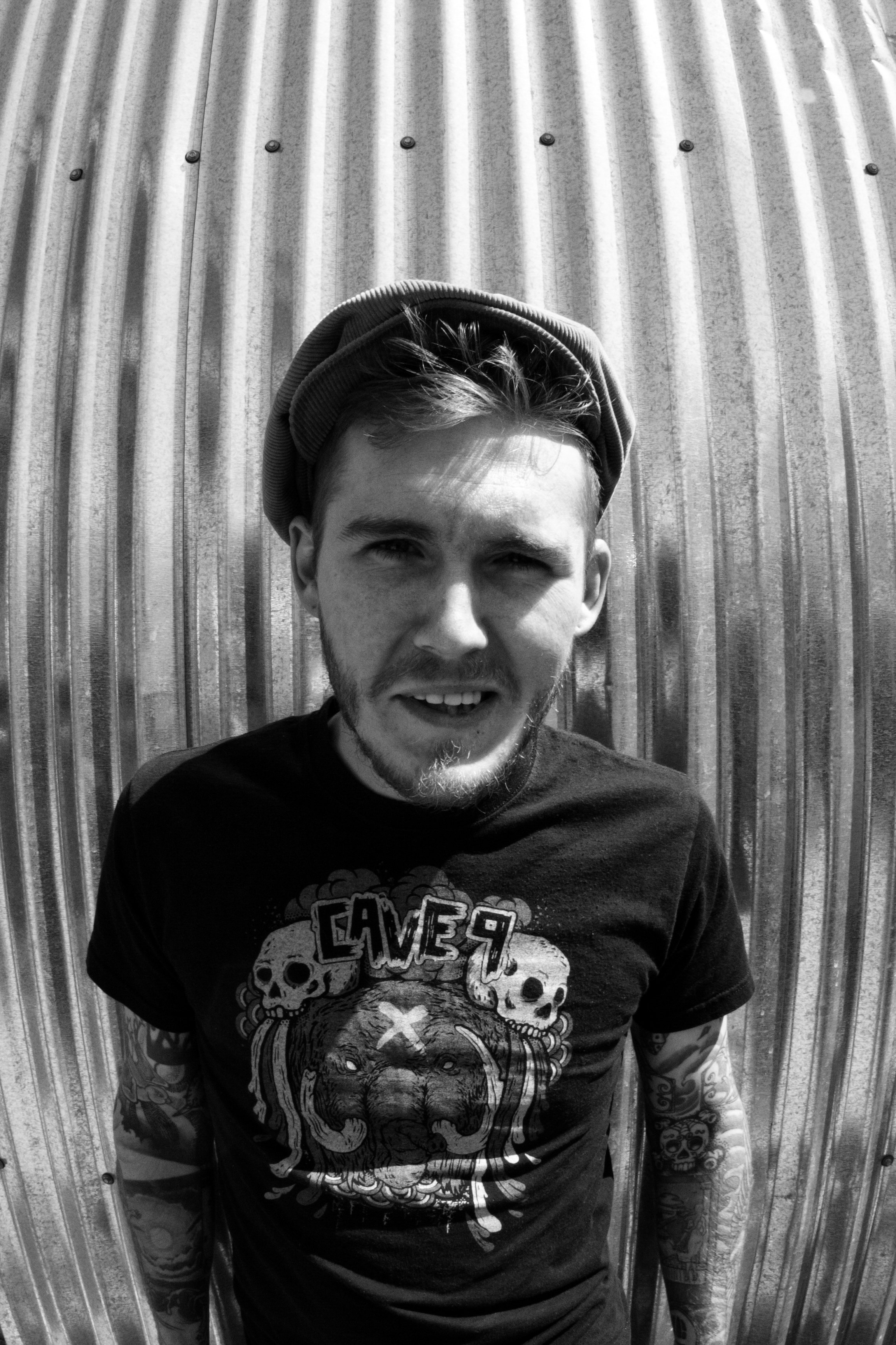 Young man with tattoos on arms wearing a graphic T-shirt, smiling, standing against a corrugated metal background.