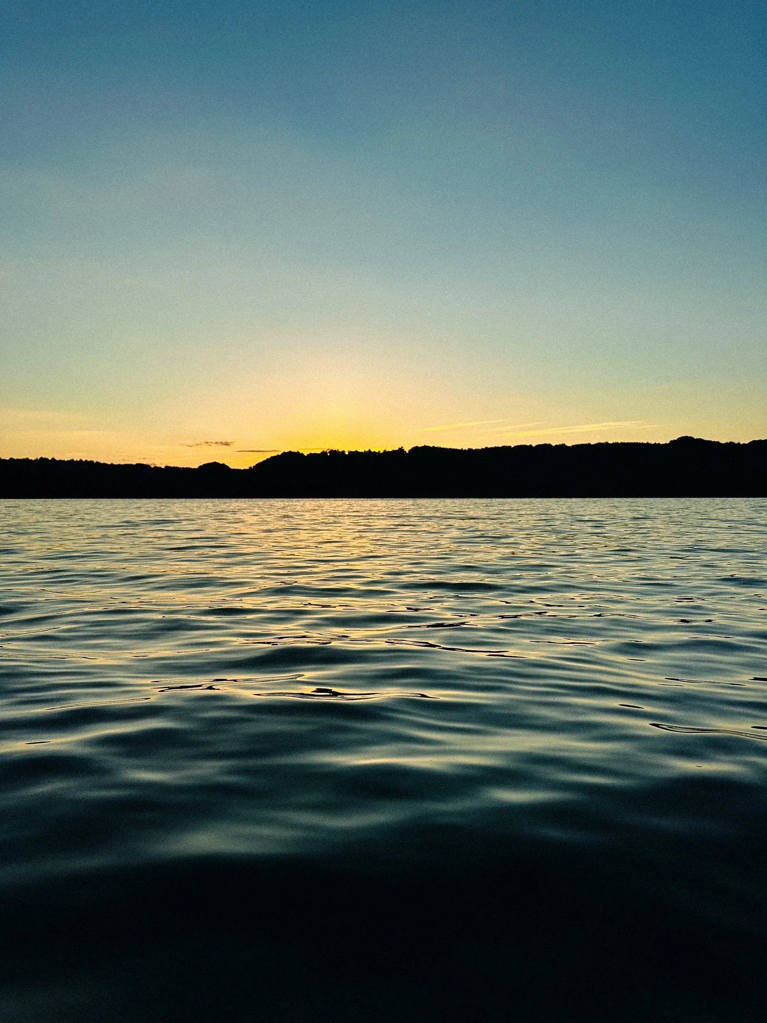 Sunset over a body of water with a silhouetted hilly landmass in the distance and ripples in the water.