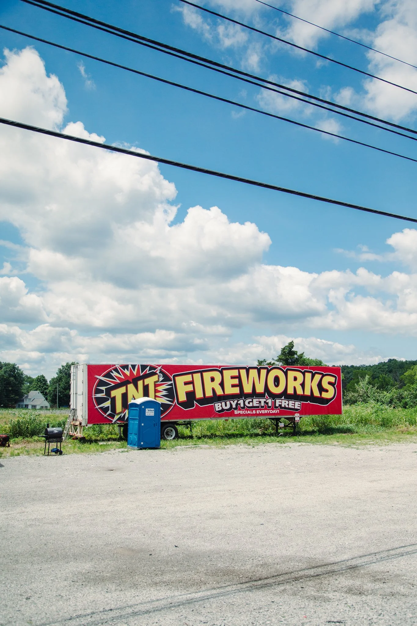A large red billboard advertising fireworks with the text "TNT Fireworks" and "Buy 1 Get 1 Free" in bold yellow and black lettering, set in an open area with a gravel ground, green trees in the background, and a blue portable toilet in front of the b