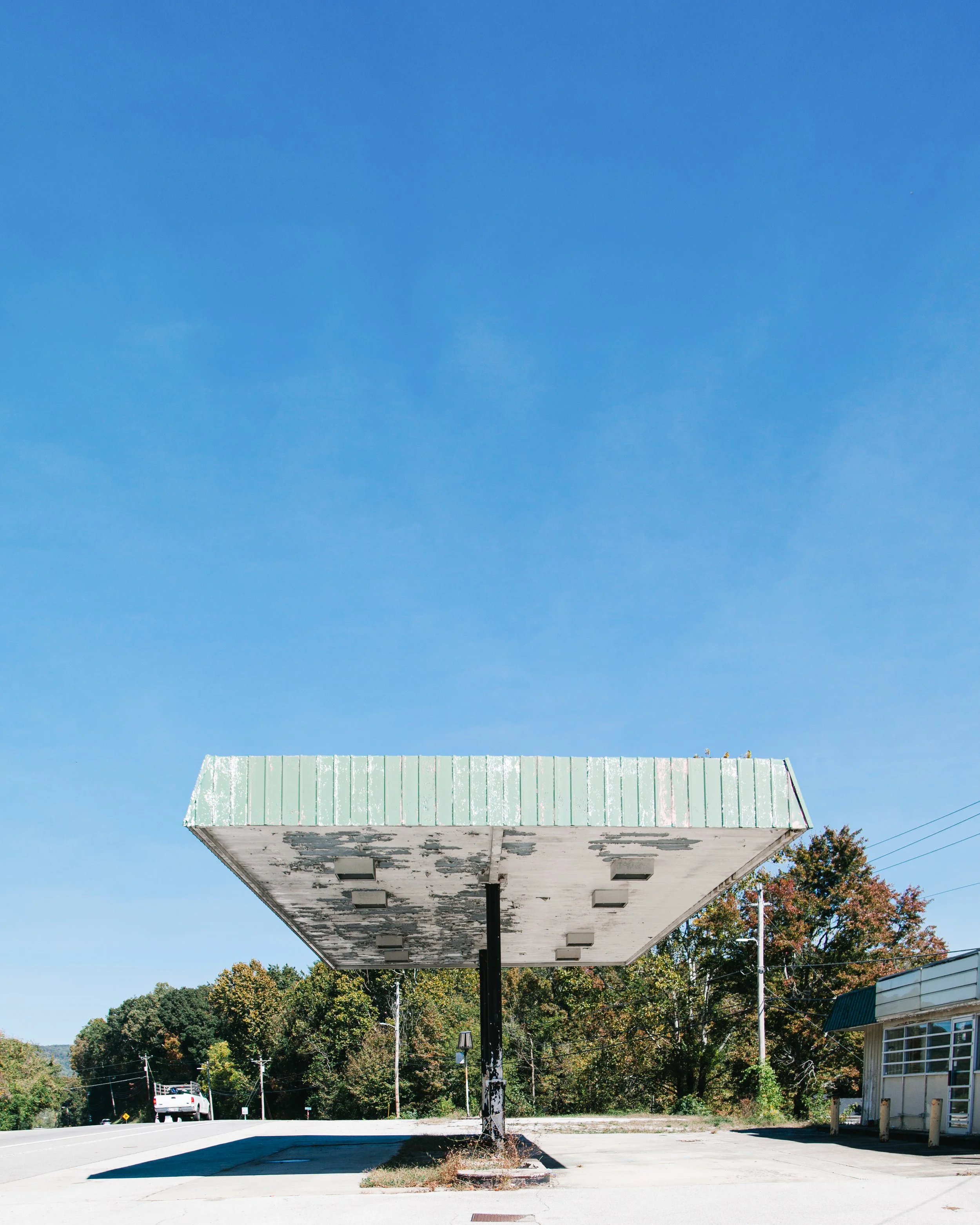 An old, empty gas station canopy with a peeling green roof, supported by a single black pole, extending over a concrete area, with trees and a small building nearby.