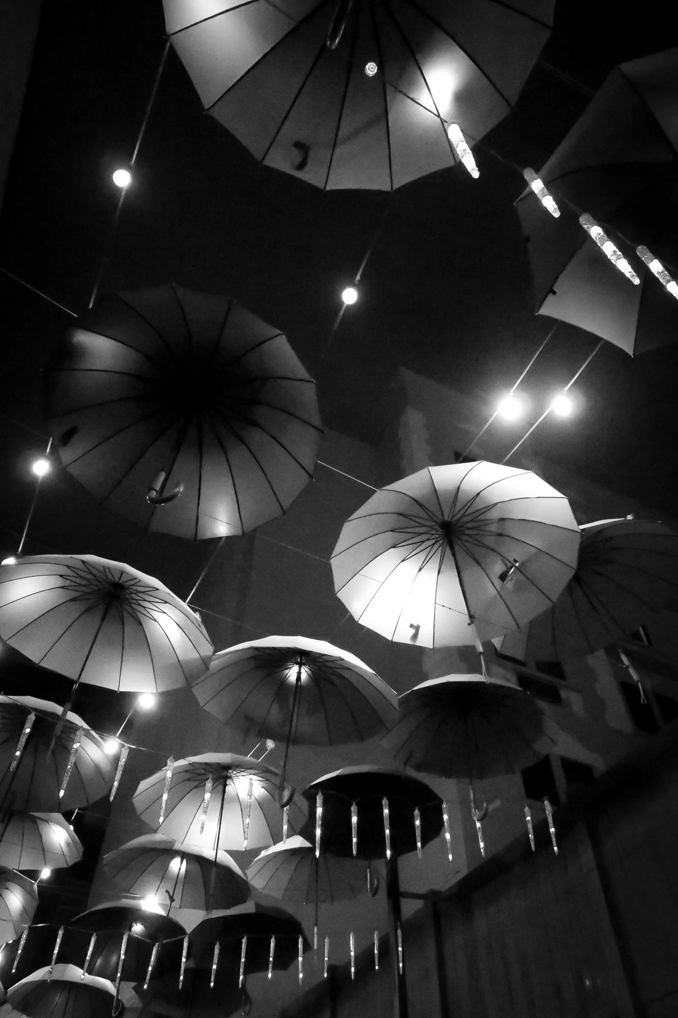 Ceiling decorated with numerous umbrellas hanging upside down, illuminated by small lights, in black and white.